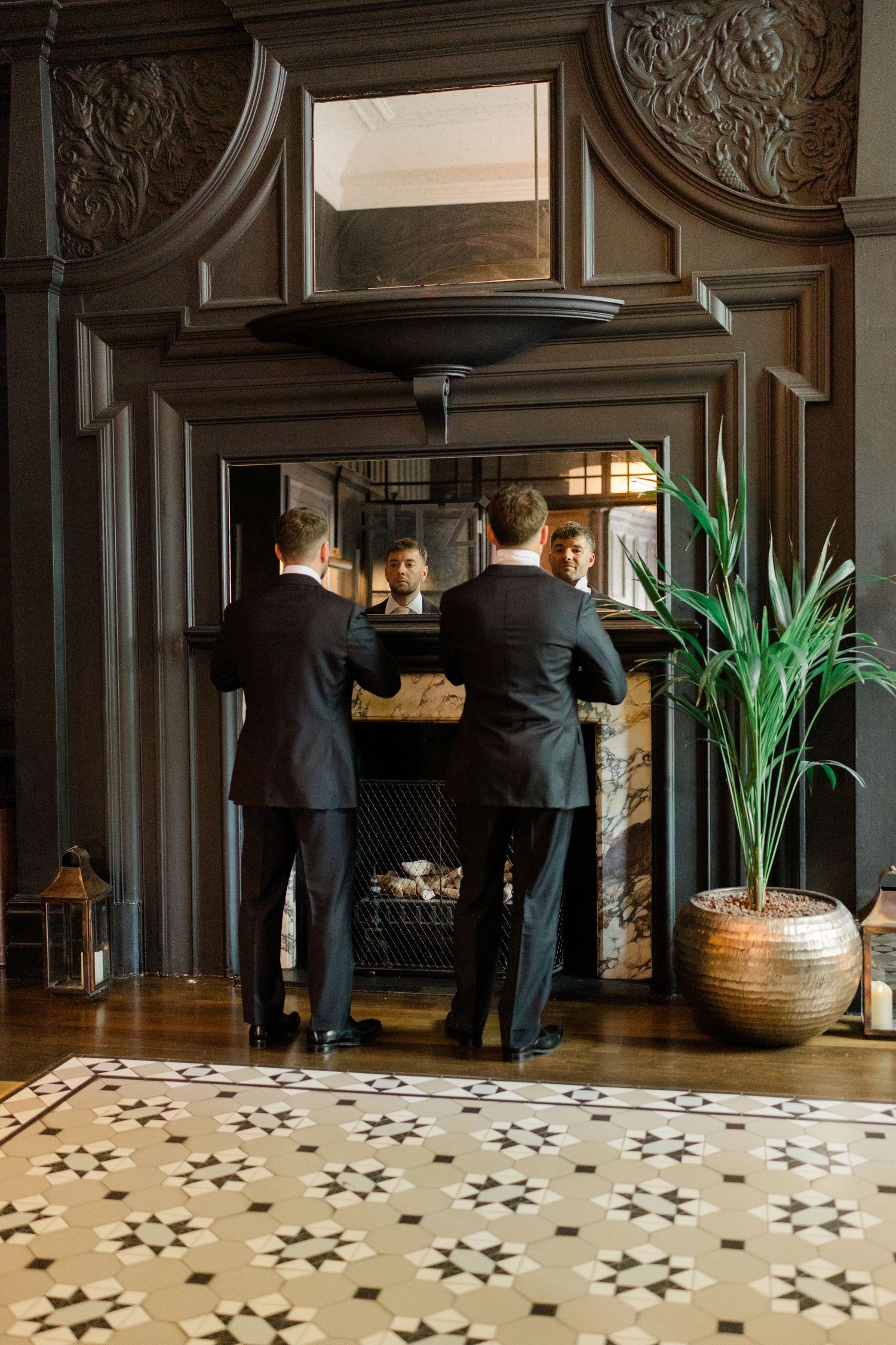 Two groomsmen in black suits stand before an ornate dark wood fireplace, their reflections visible in the mirror above.