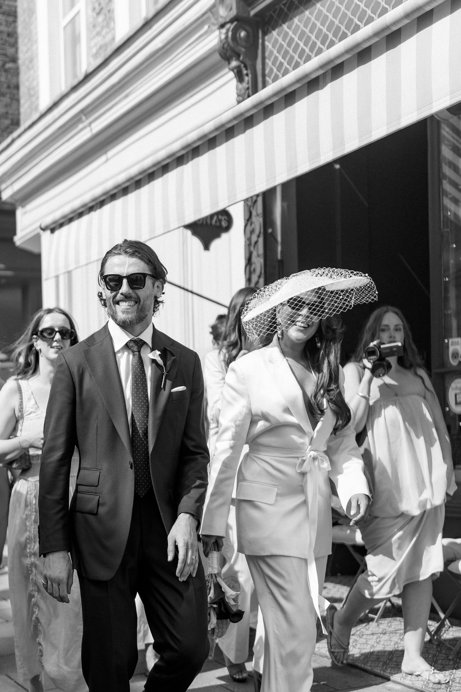 Black and white photo of smiling groom in dark suit and bride in white suit with wide-brim hat.