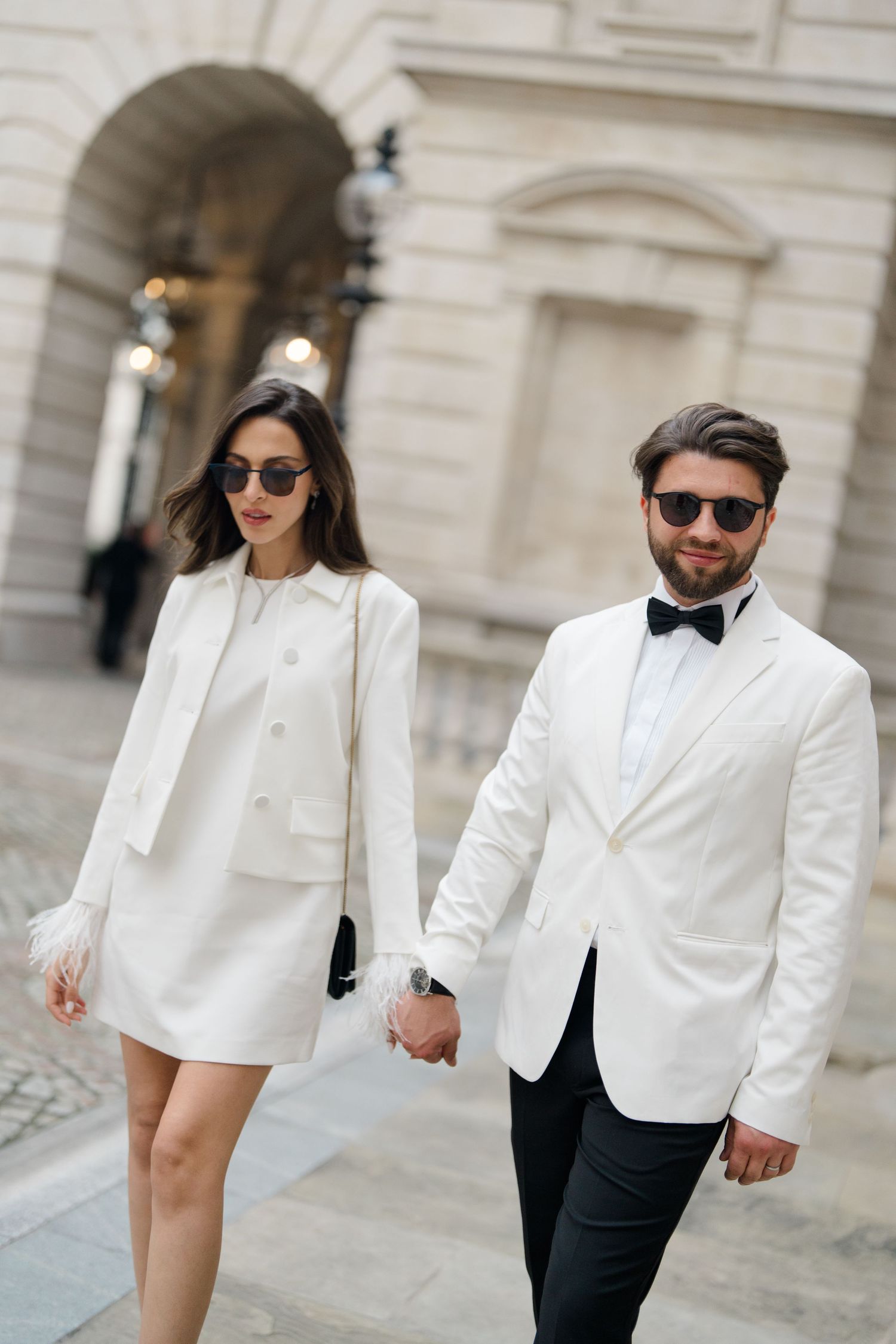 Elegant couple in matching white outfits and sunglasses holding hands outside a classic building.