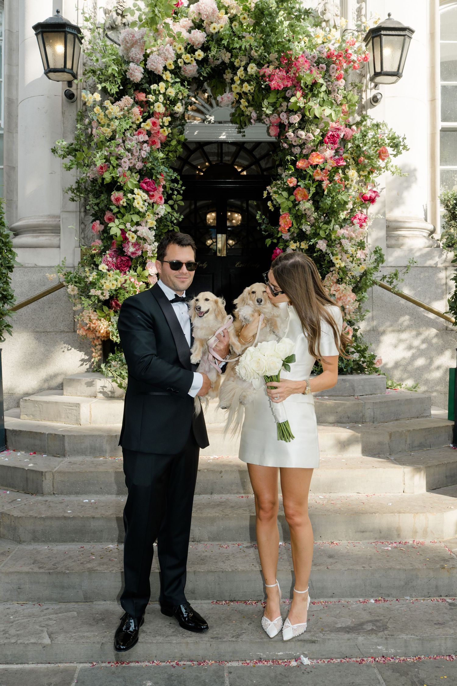 Bride and groom in sunglasses hold fluffy dogs before elaborate floral arch at venue entrance.