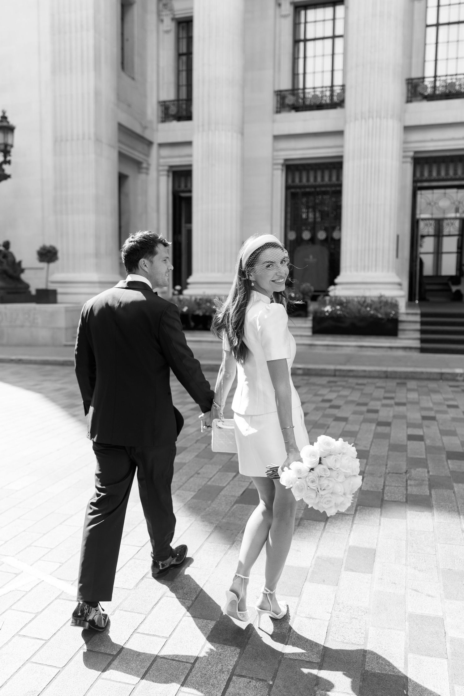 Black and white photo of bride and groom holding hands walking past grand columned building on wedding day.