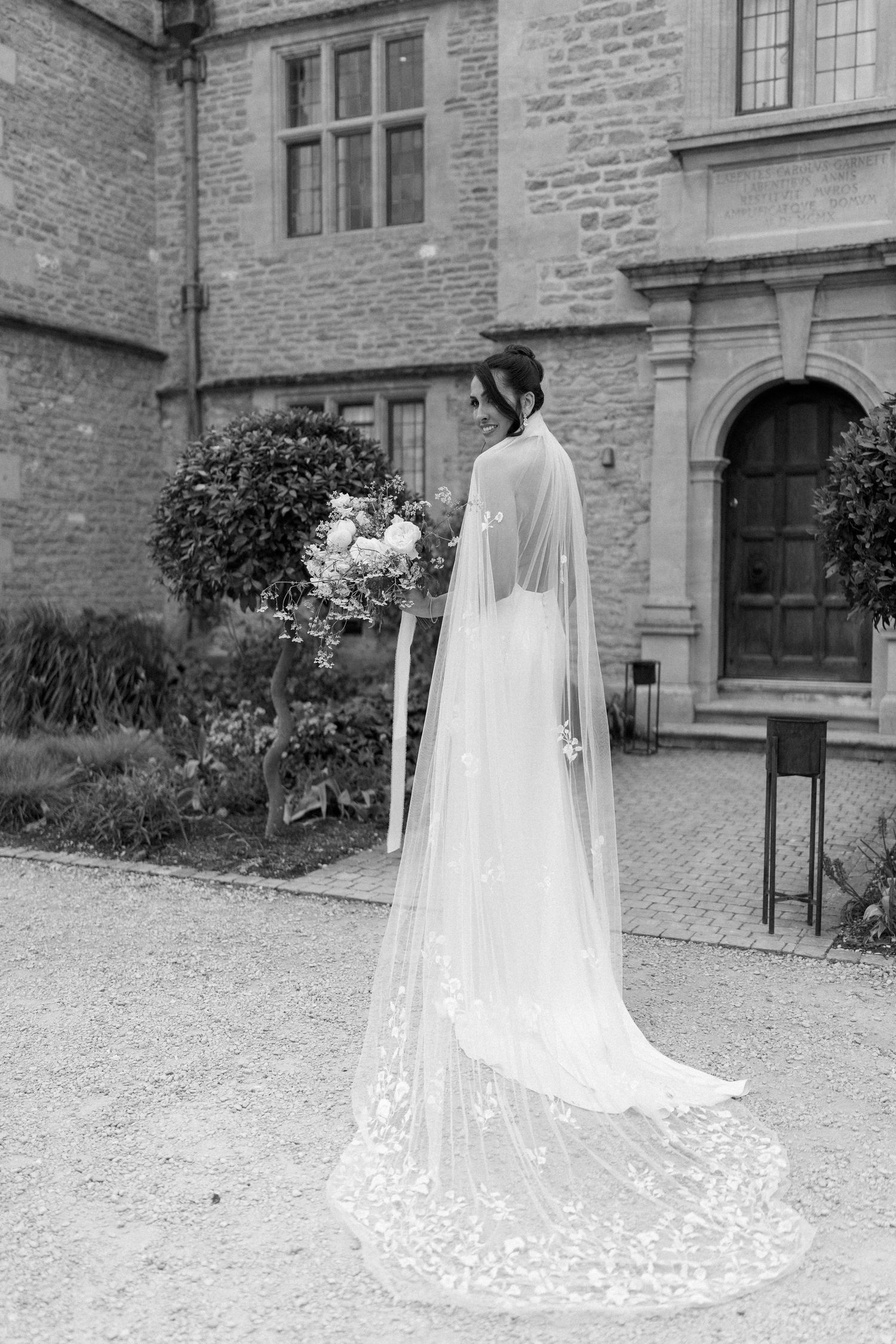 Bride holding bouquet in a sleek wedding dress with a lace cathedral veil outside a stone manor.