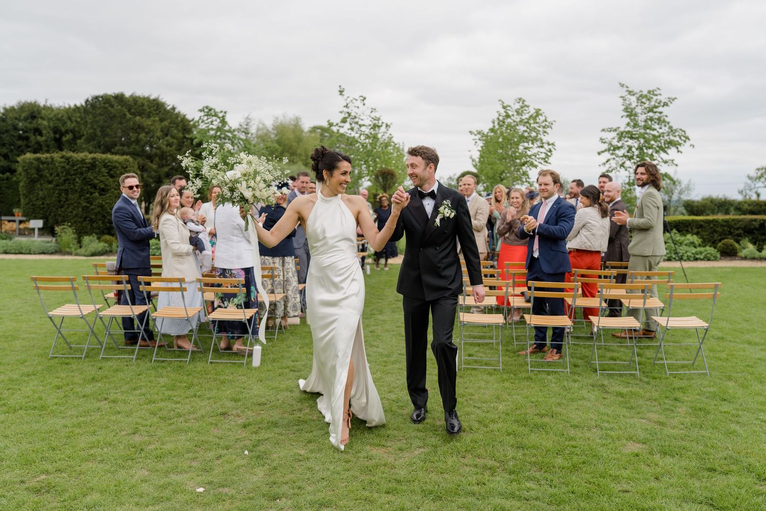 Newlywed couple walks hand in hand past cheering guests at outdoor garden wedding.