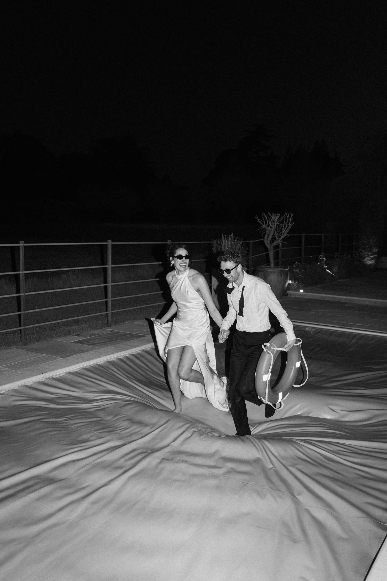 Bride and groom in sunglasses jumping on inflatable at night in fun black and white wedding photo.