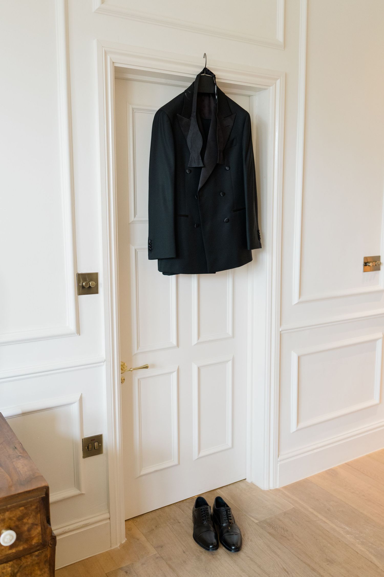 Black double-breasted tuxedo jacket hanging on a white paneled door above polished black dress shoes on hardwood floor.