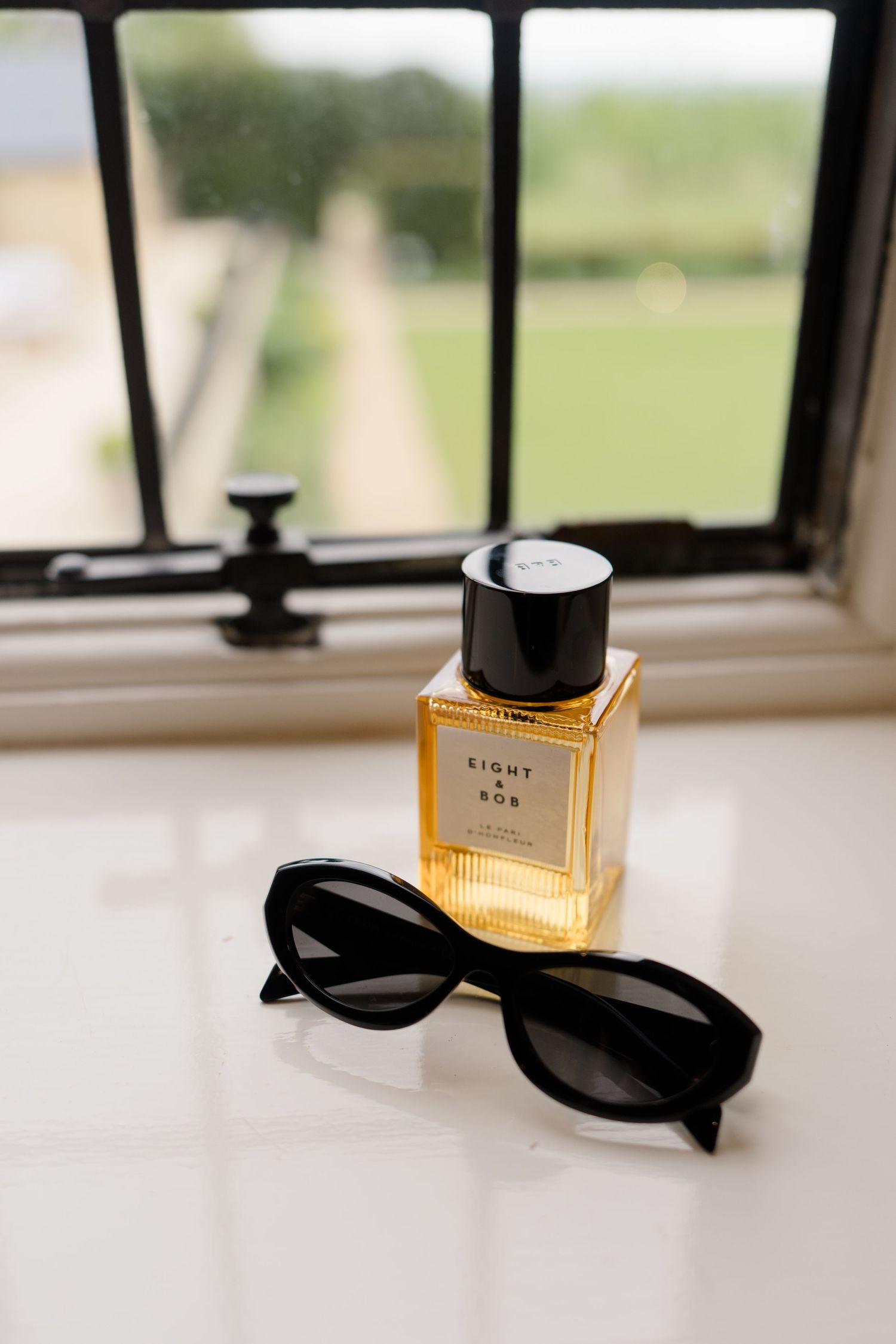 Eight and Bob perfume bottle with black sunglasses on a white windowsill overlooking a garden.