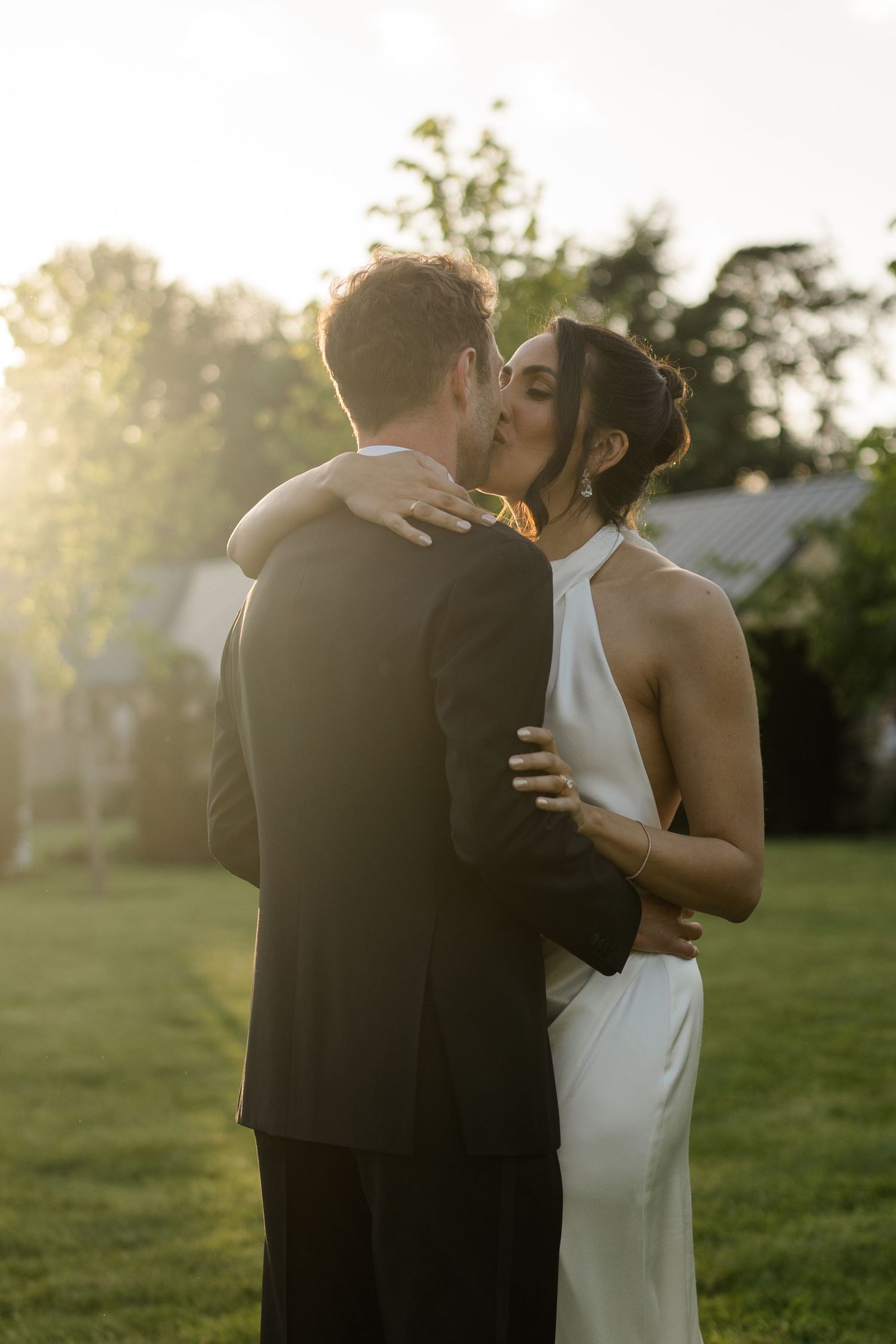 Bride and groom share a romantic kiss outdoors in golden sunset light at their wedding.