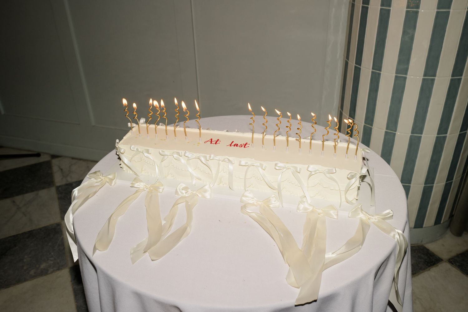 Wedding cake decorated with lit candles and white ribbon streamers on round table with marble floor background.