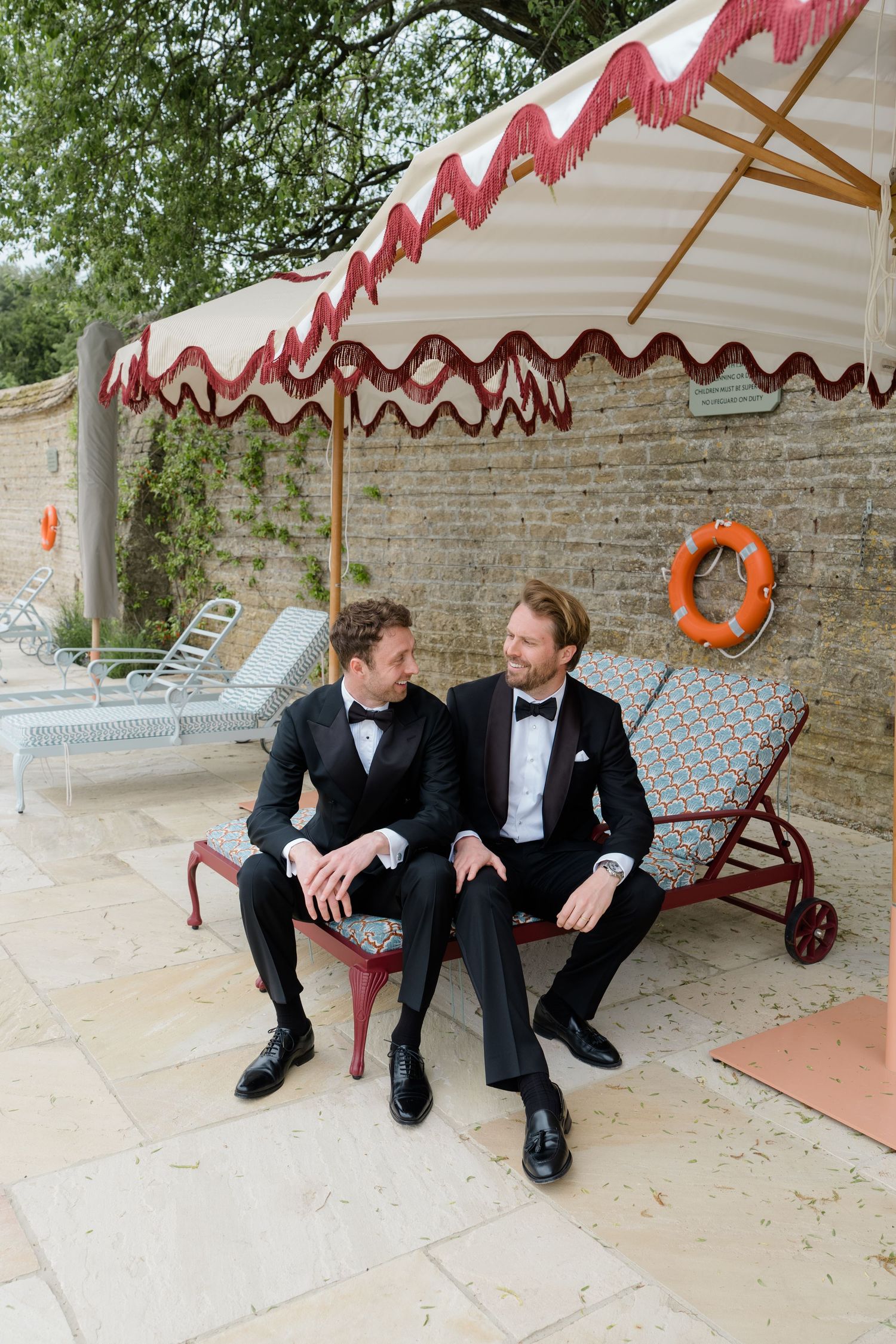 Two grooms in black tuxedos sit together on a lounge chair by a poolside umbrella.