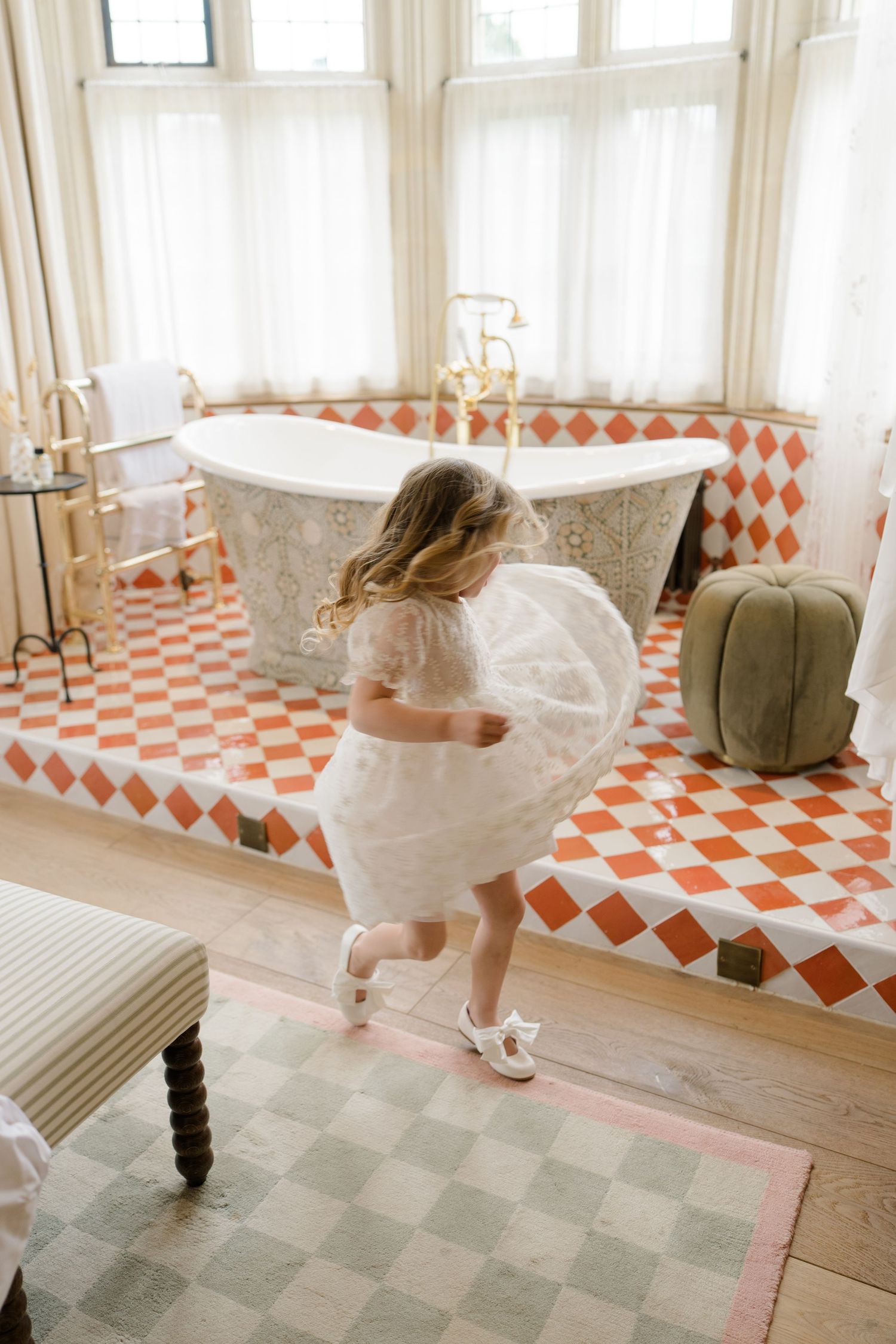 Flower girl in white tutu dress twirling in sunlit bathroom with clawfoot tub.