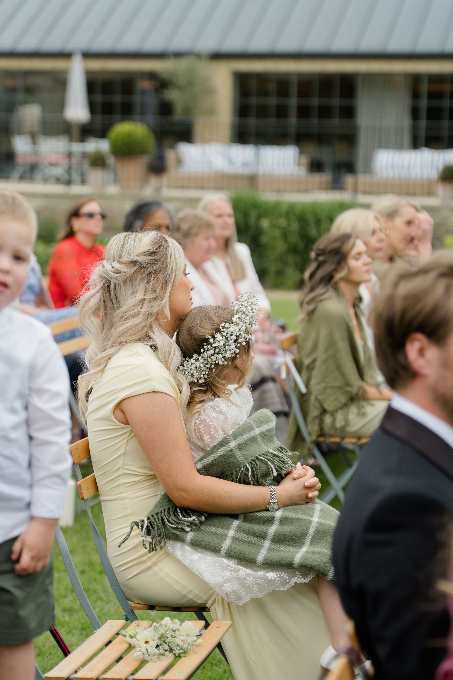 A wedding guest in yellow holds a sleeping flower girl with a floral crown during an outdoor ceremony.