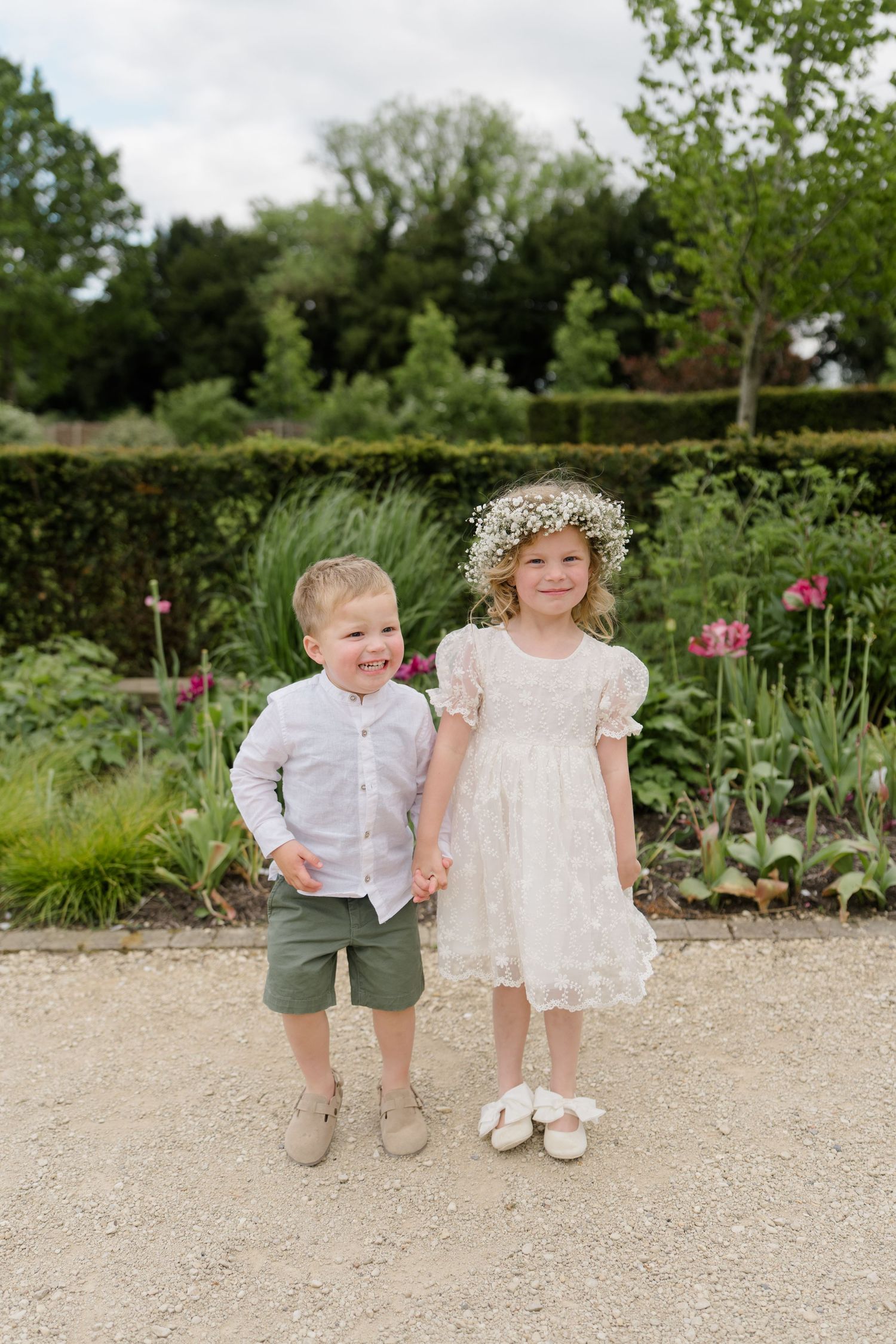 Adorable flower girl in white lace dress with floral crown holds hands with young ring bearer outdoors.