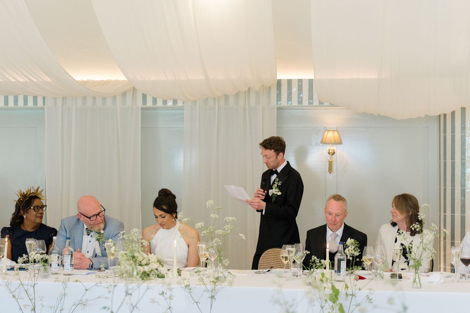 Groom delivers speech at wedding reception head table in elegant striped marquee venue.