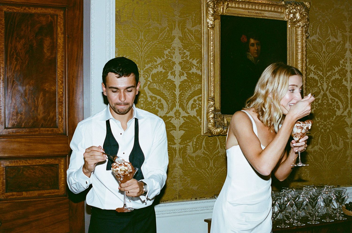 Couple at wedding reception, man eating dessert and woman drinking champagne, gold wallpaper background.
