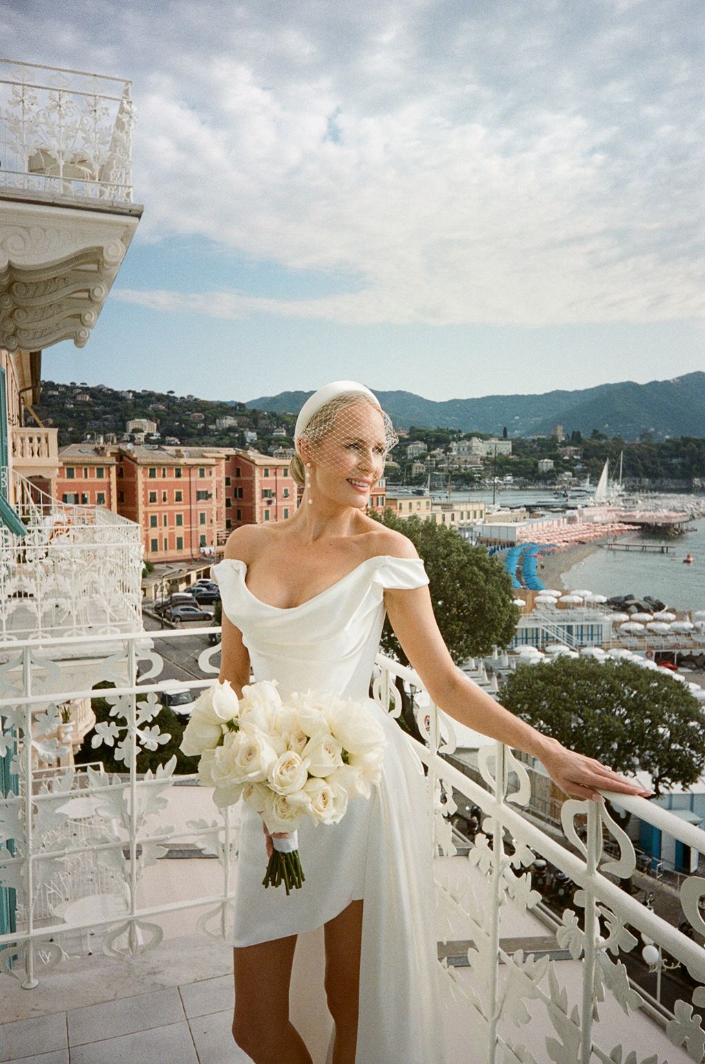 Elegant bride in white off-shoulder gown holding bouquet on balcony with coastal town view.