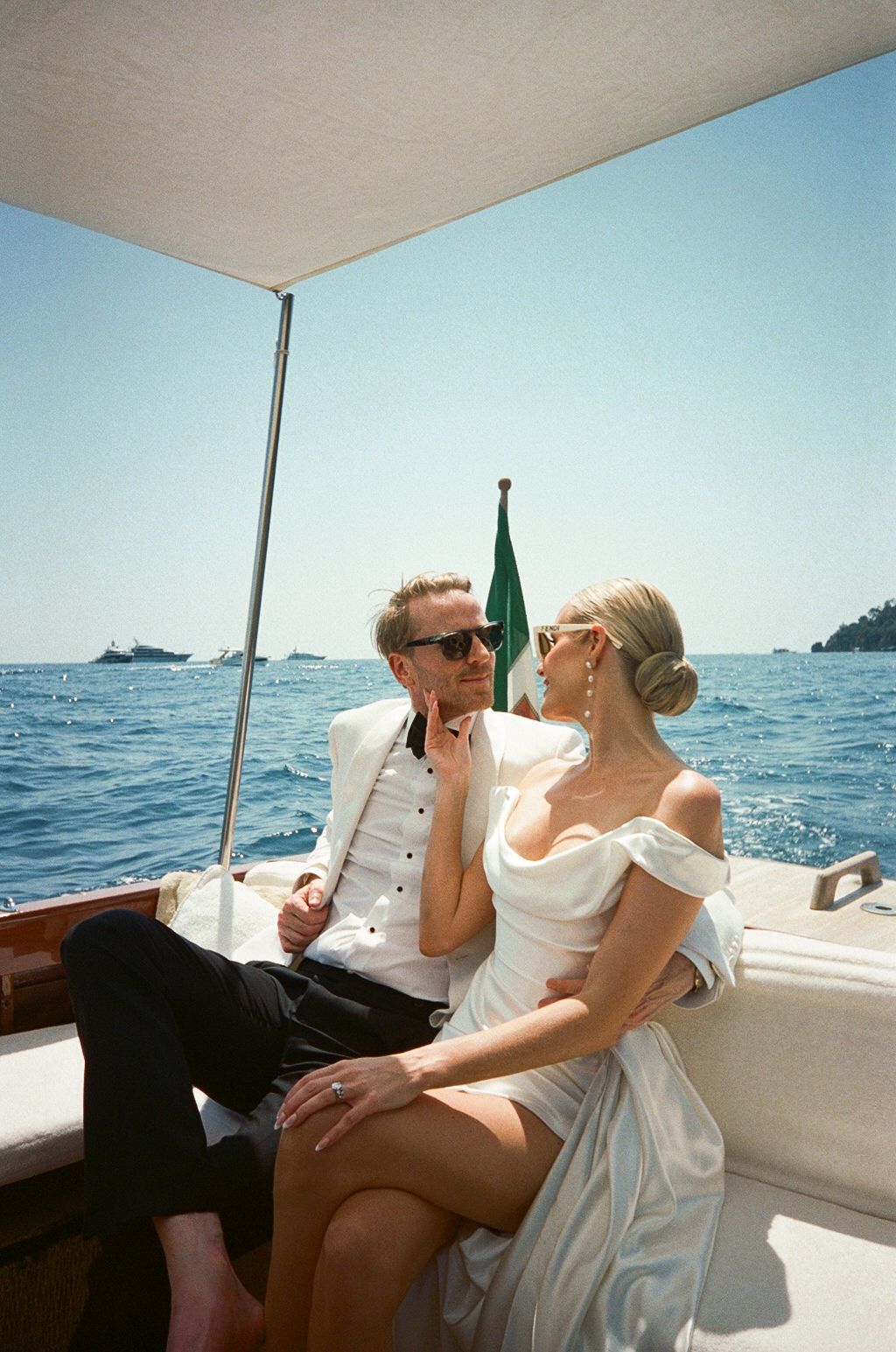 Newlyweds in wedding attire sit closely on a wooden boat with the Italian flag and blue sea in the background.
