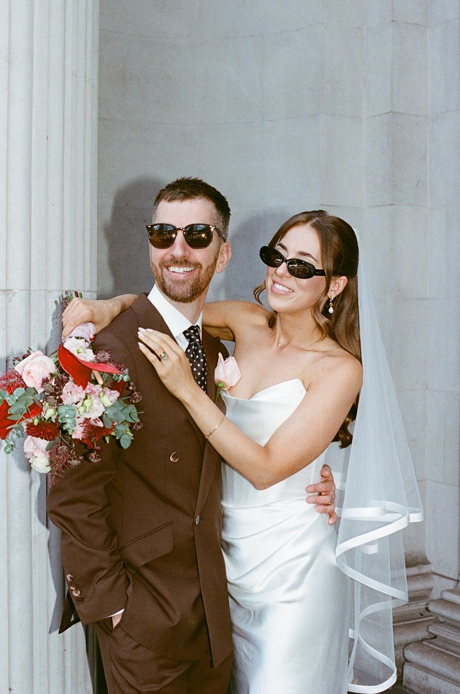 Smiling newlyweds in sunglasses pose together holding a red and pink bridal bouquet.