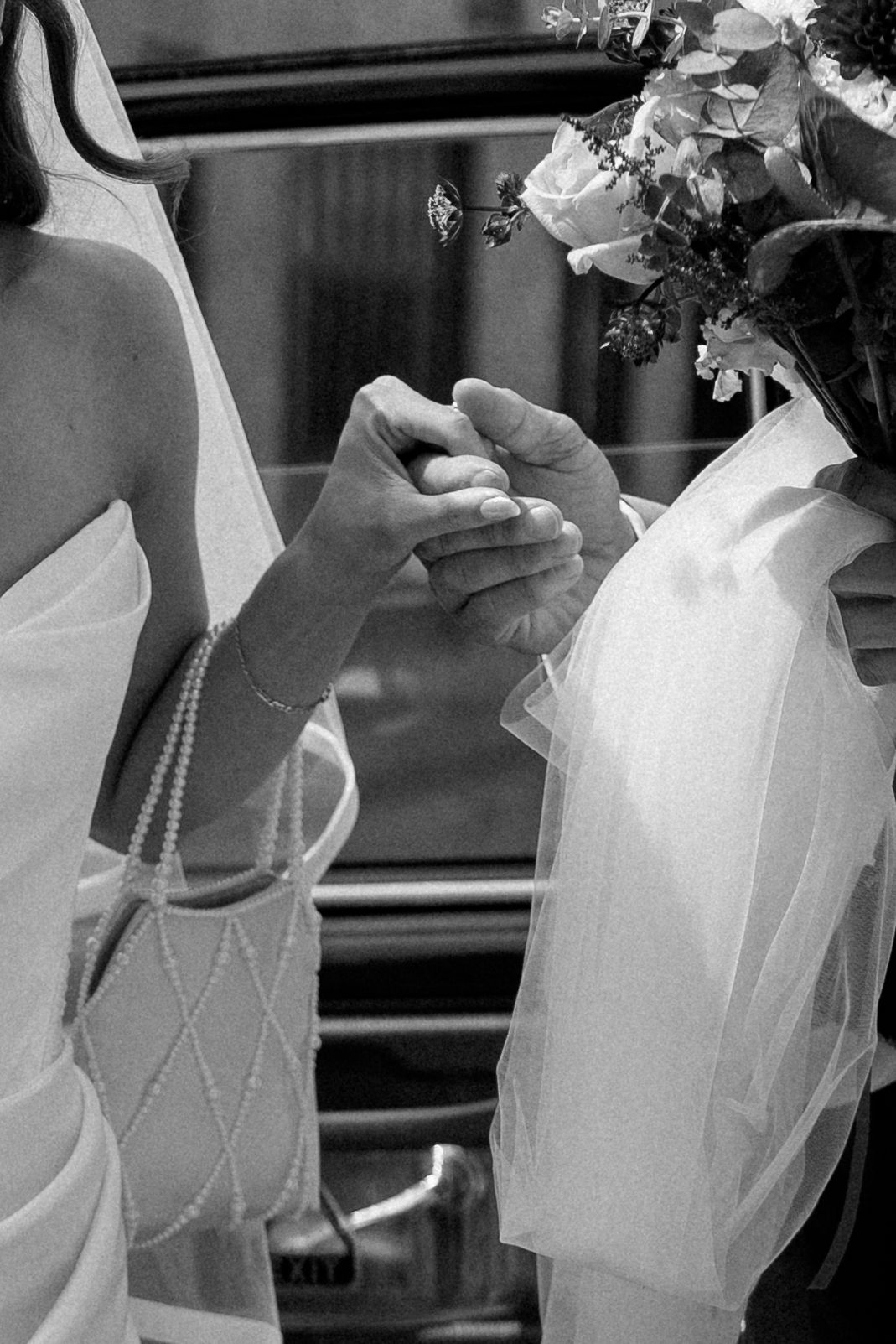 Black and white photo of two brides holding hands, one wearing pearl bracelet, exchanging rings at wedding ceremony.