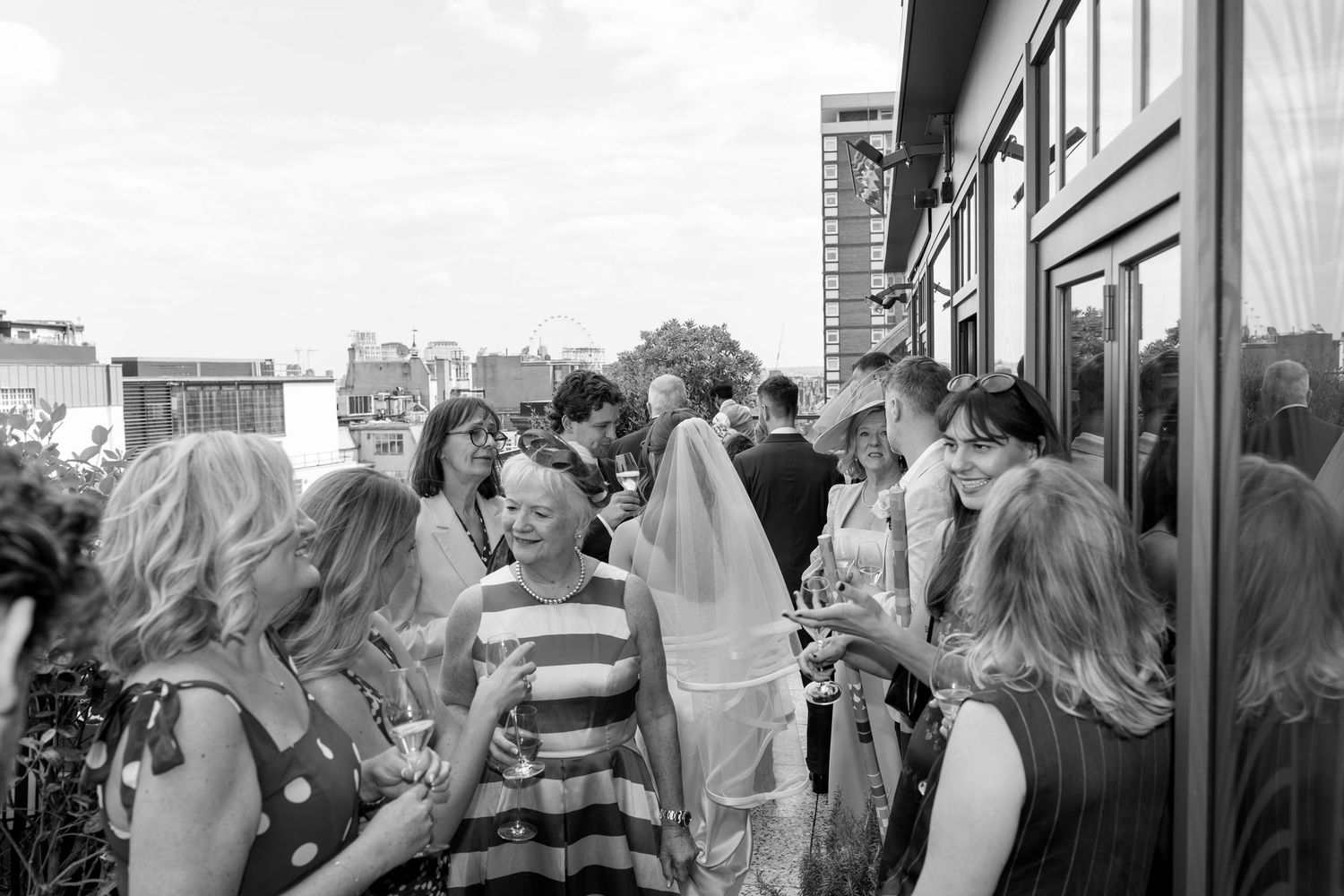 Wedding guests mingle on a rooftop terrace with the bride visible in her veil among the crowd.