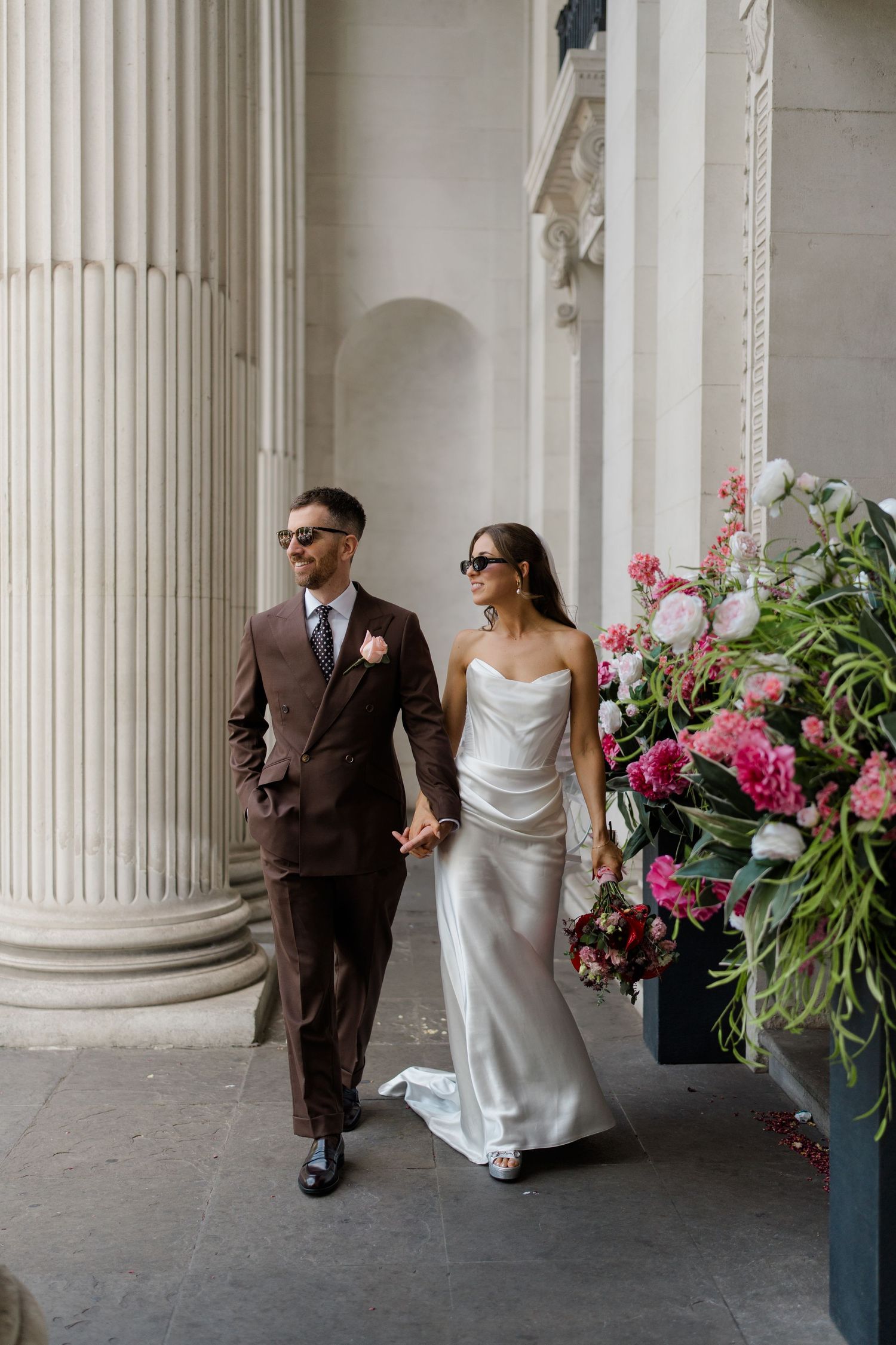 Bride and groom walk hand in hand past large stone columns and pink floral arrangements after their wedding.