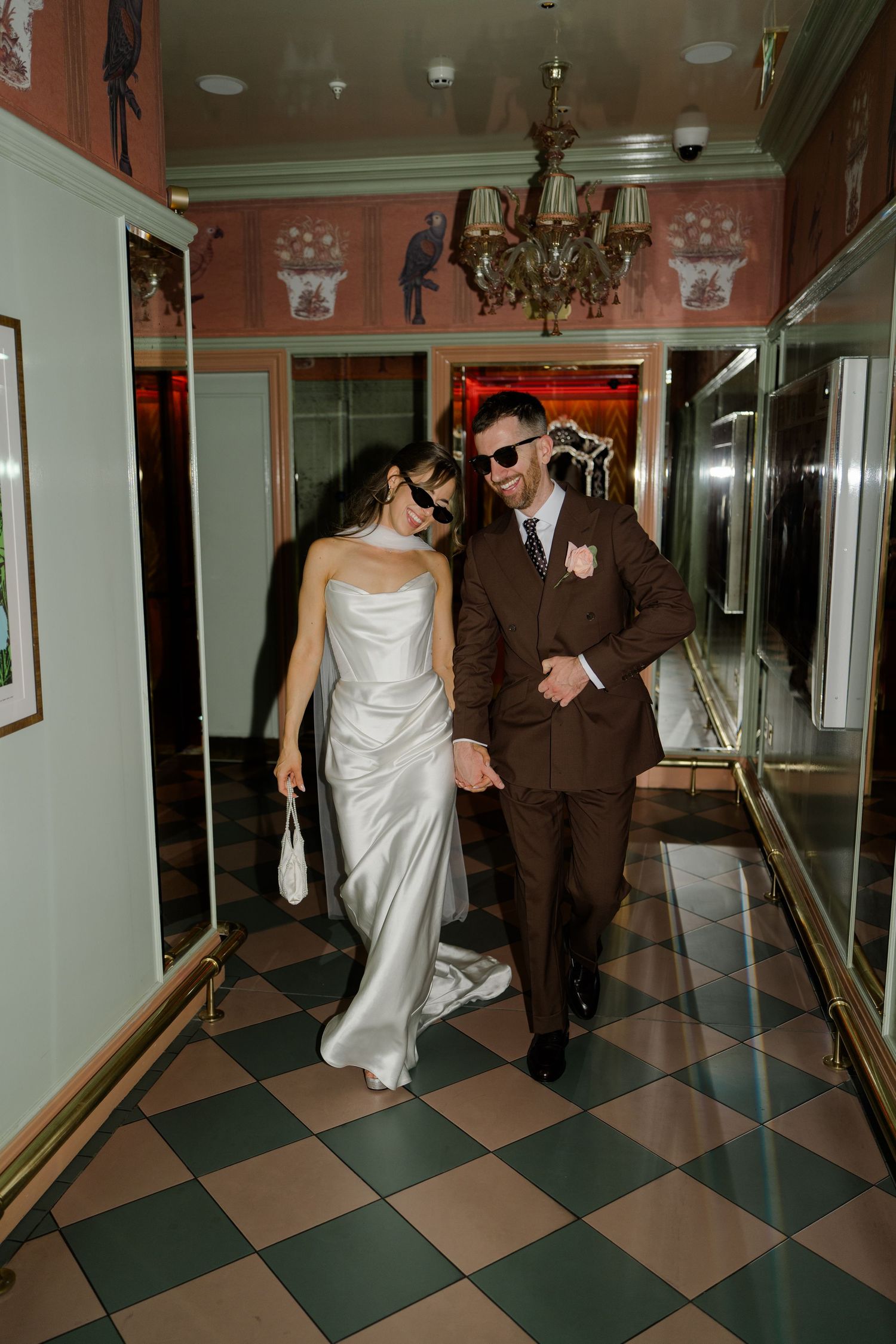 Bride and groom in sunglasses walking through ornate hotel corridor with checkered floor.