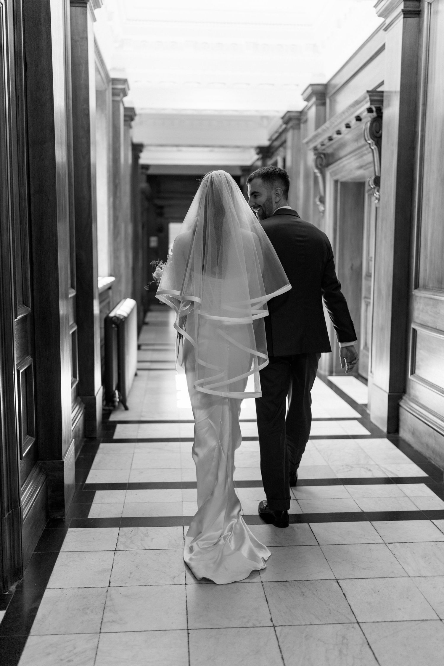 Black and white rear view of bride and groom walking through grand columned hallway after ceremony.