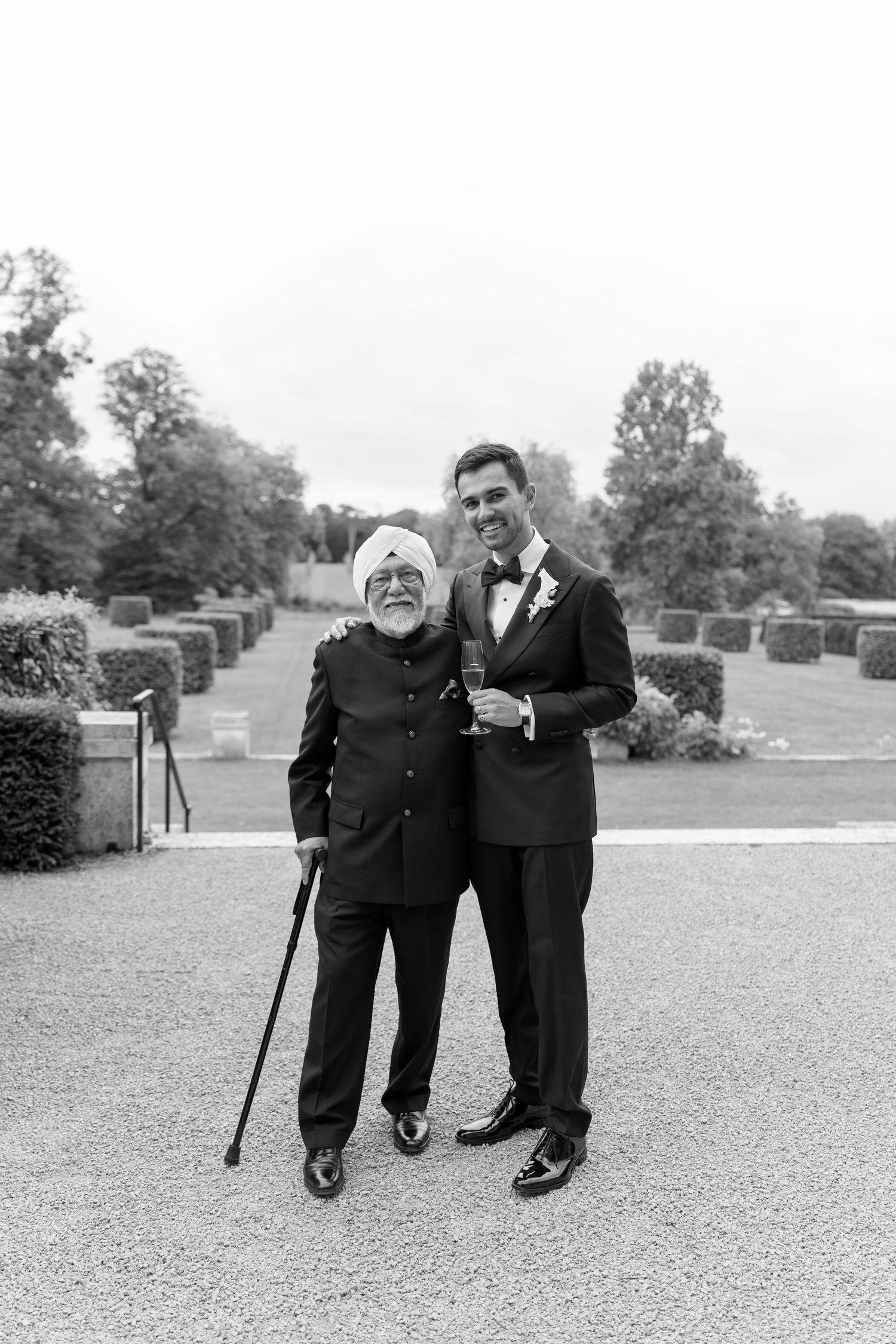 An elderly Sikh man with a cane poses with a groom in a tuxedo at an outdoor wedding venue.
