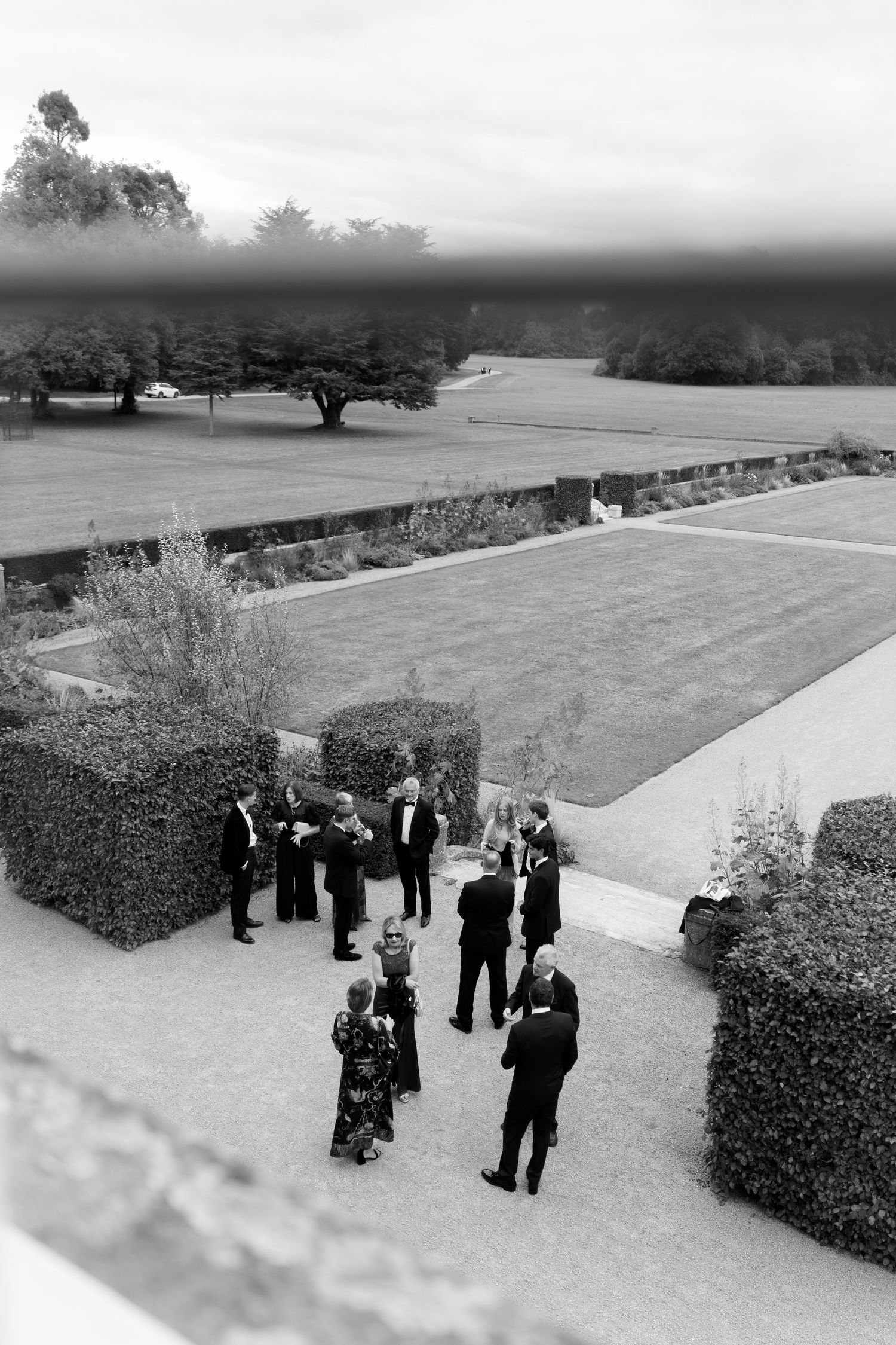 Aerial black and white photo of elegantly dressed guests mingling on a garden terrace at a formal outdoor event.