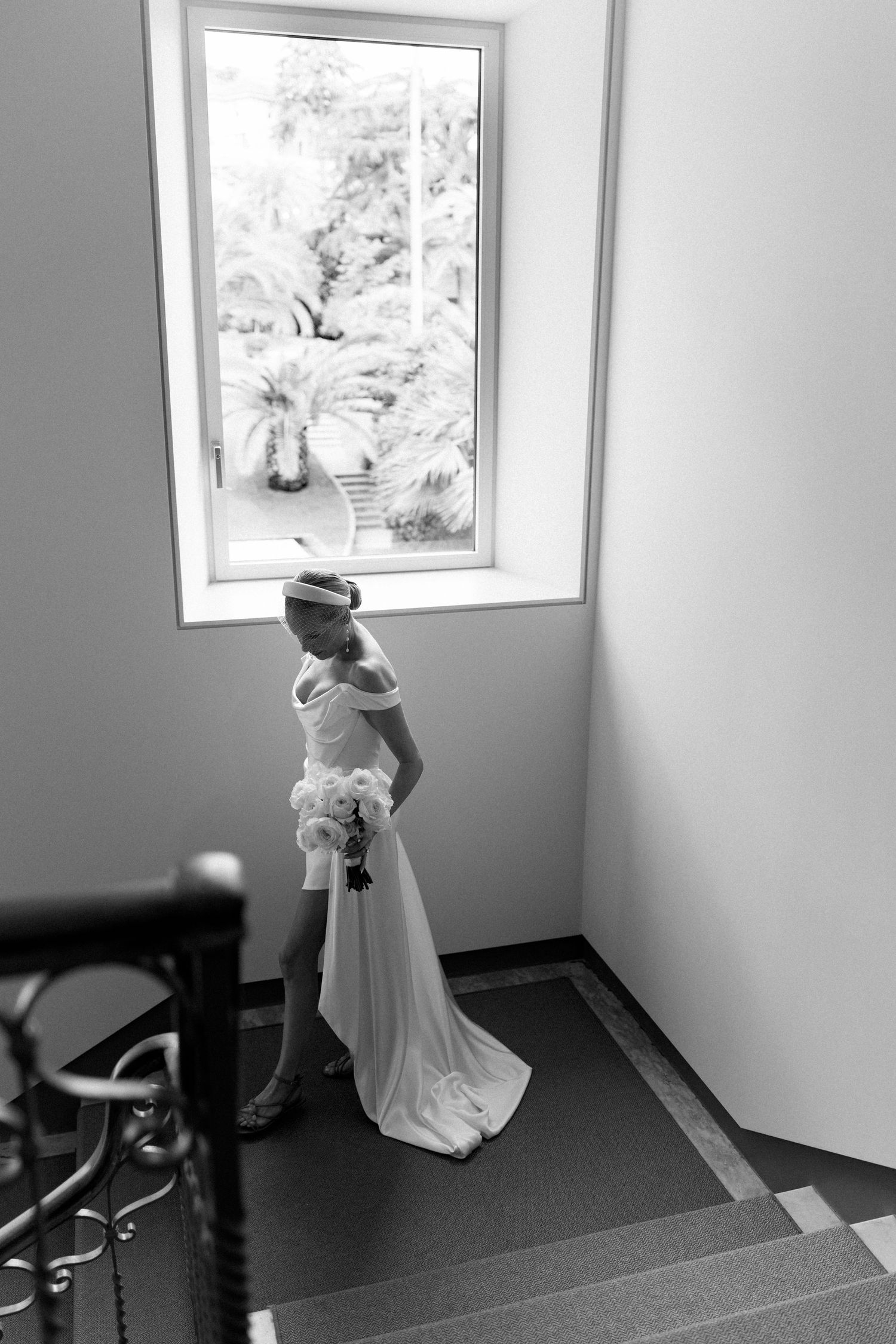 Bride in white off-shoulder gown holding bouquet stands by window in black and white wedding photo.