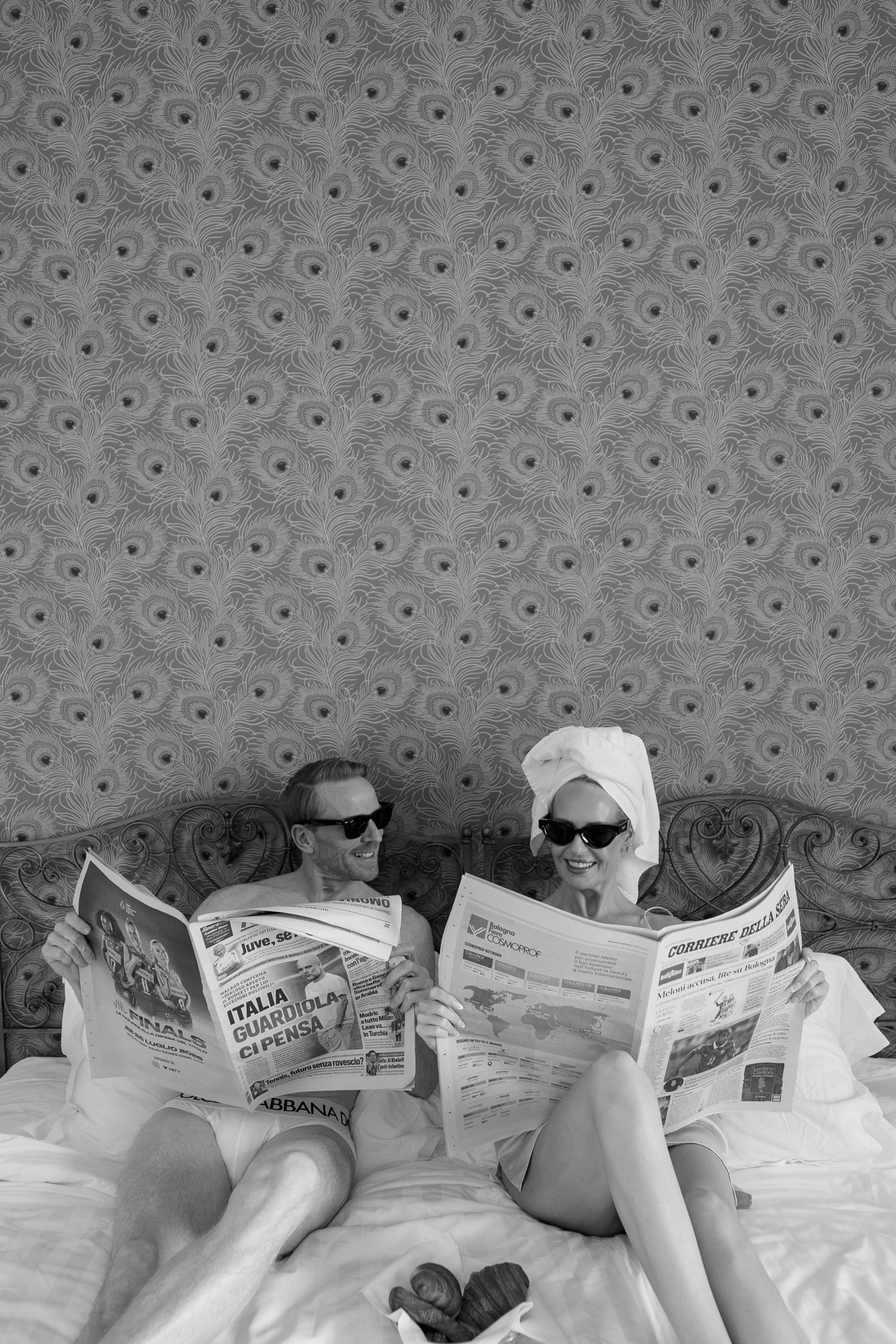 Couple in robes and towel turbans wearing sunglasses reading Italian newspapers in bed, black and white photo.