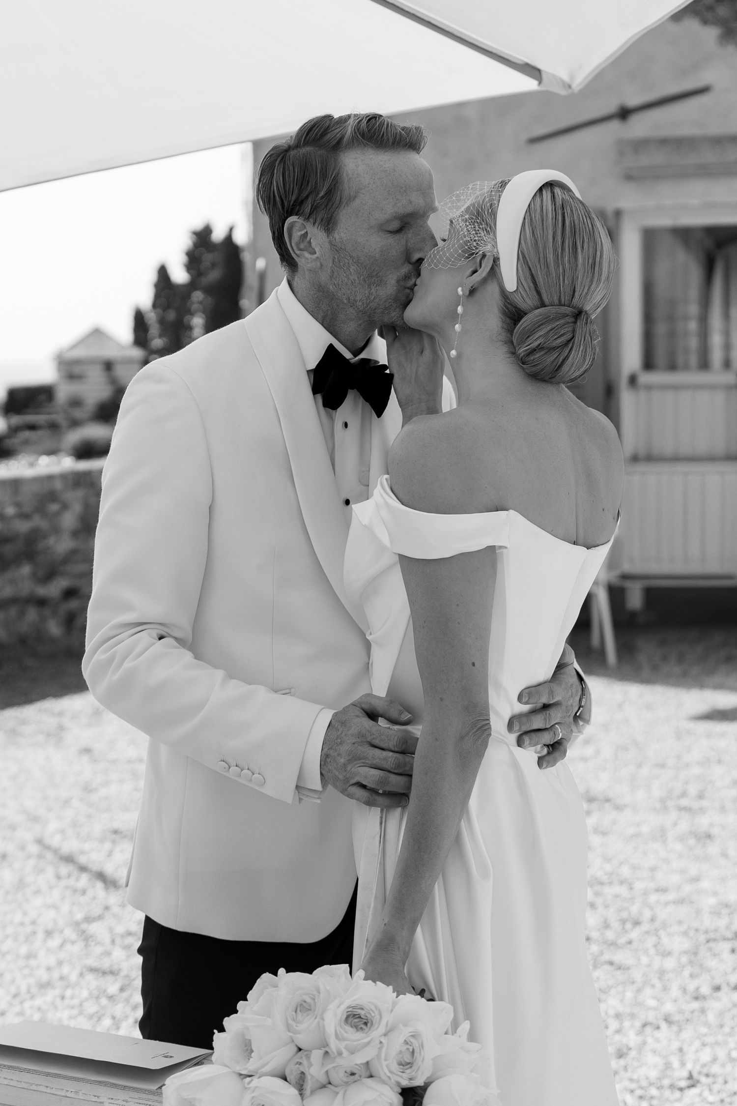 Black and white photo of bride and groom sharing first kiss at outdoor wedding ceremony, groom in white tuxedo jacket.