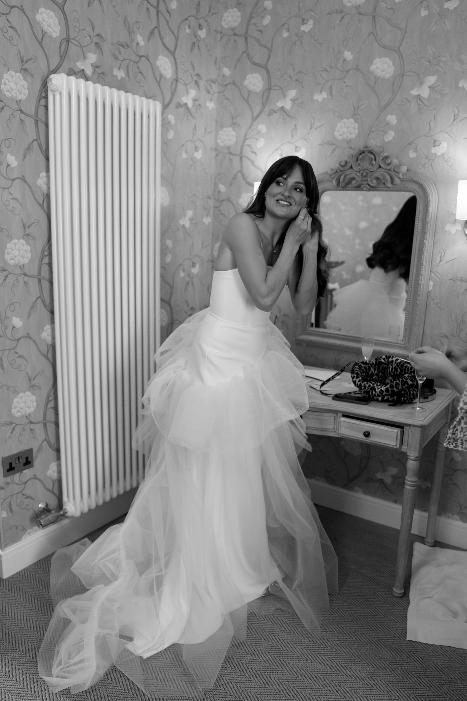 A bride in a flowing white gown poses by a vintage mirror and radiator against floral wallpaper.