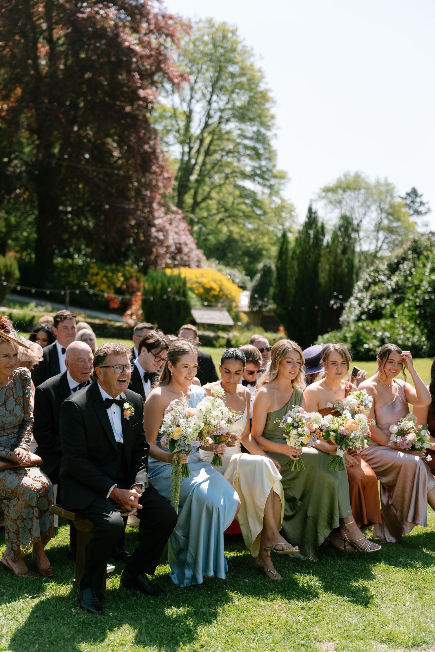 Wedding party relaxes on grass in coordinated attire during outdoor celebration.