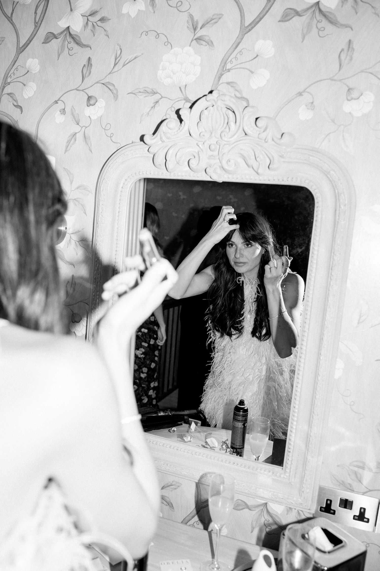 A vintage-style black and white photo showing someone applying makeup in an ornate mirror with floral wallpaper.