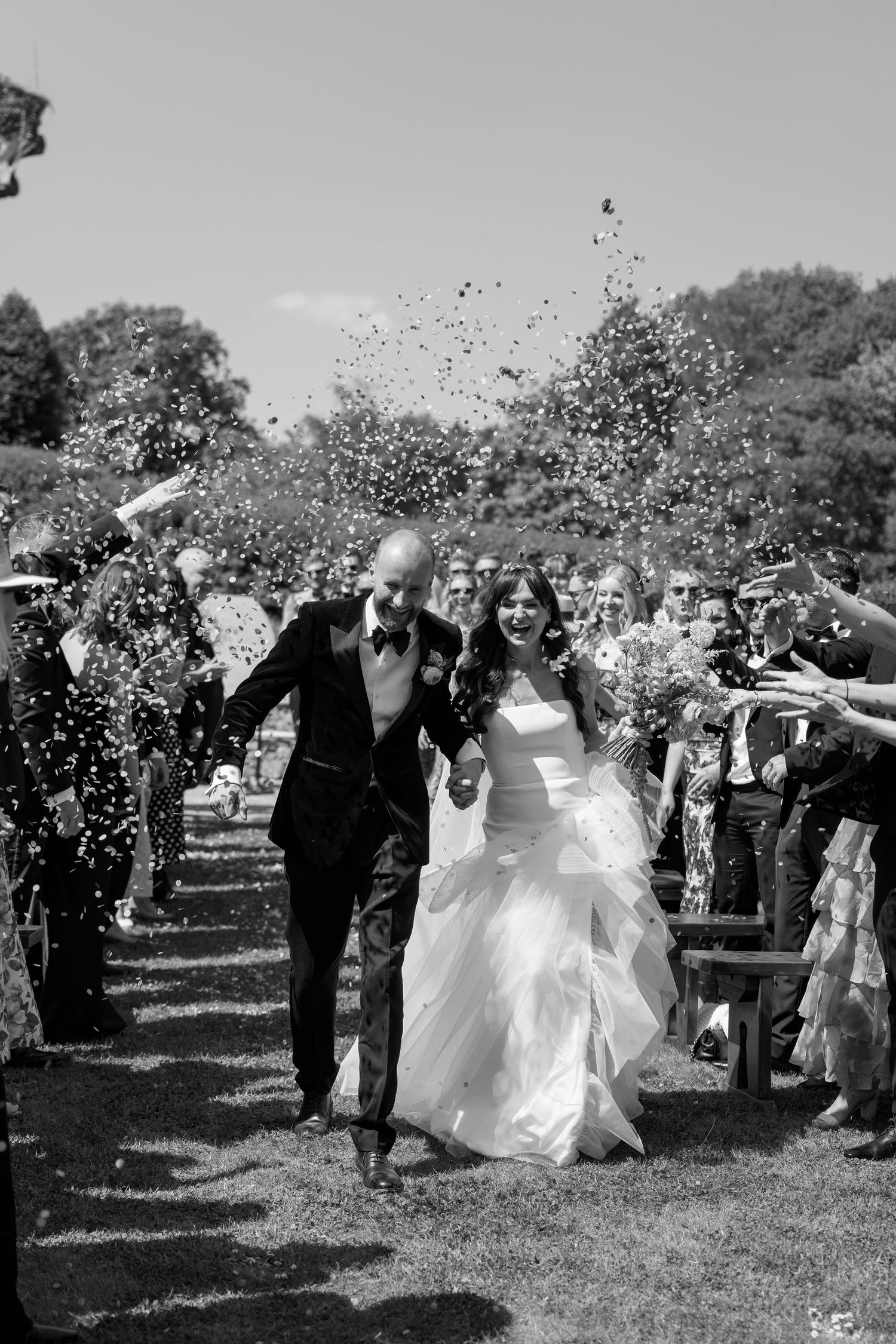Black and white photo of wedding guests showering the couple with confetti in a garden setting.