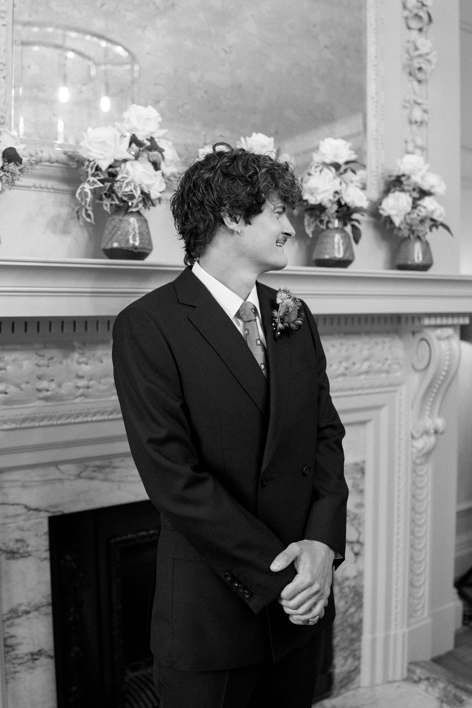 Person in formal suit standing by ornate fireplace with floral decorations.