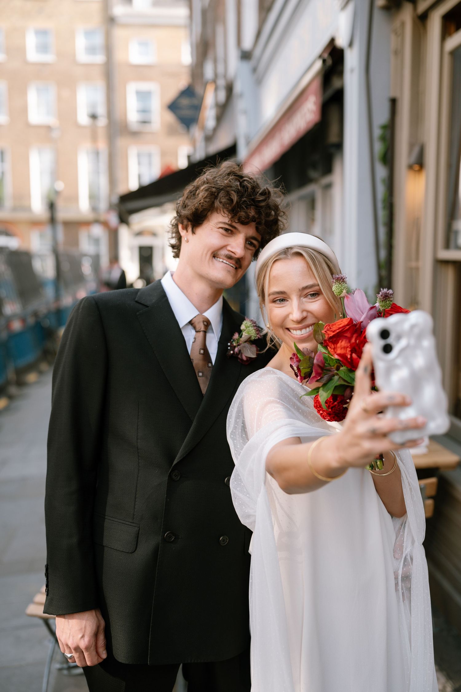 A couple takes selfies on their wedding day while holding a bouquet of red flowers on a city street.