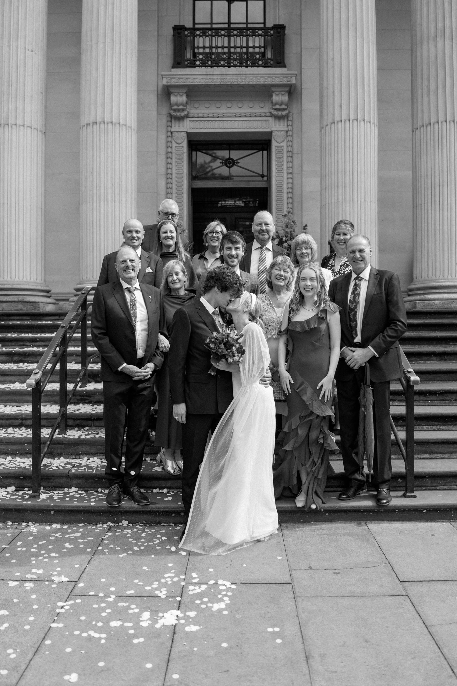Wedding party gathered on grand stone steps of historic building with classical columns.