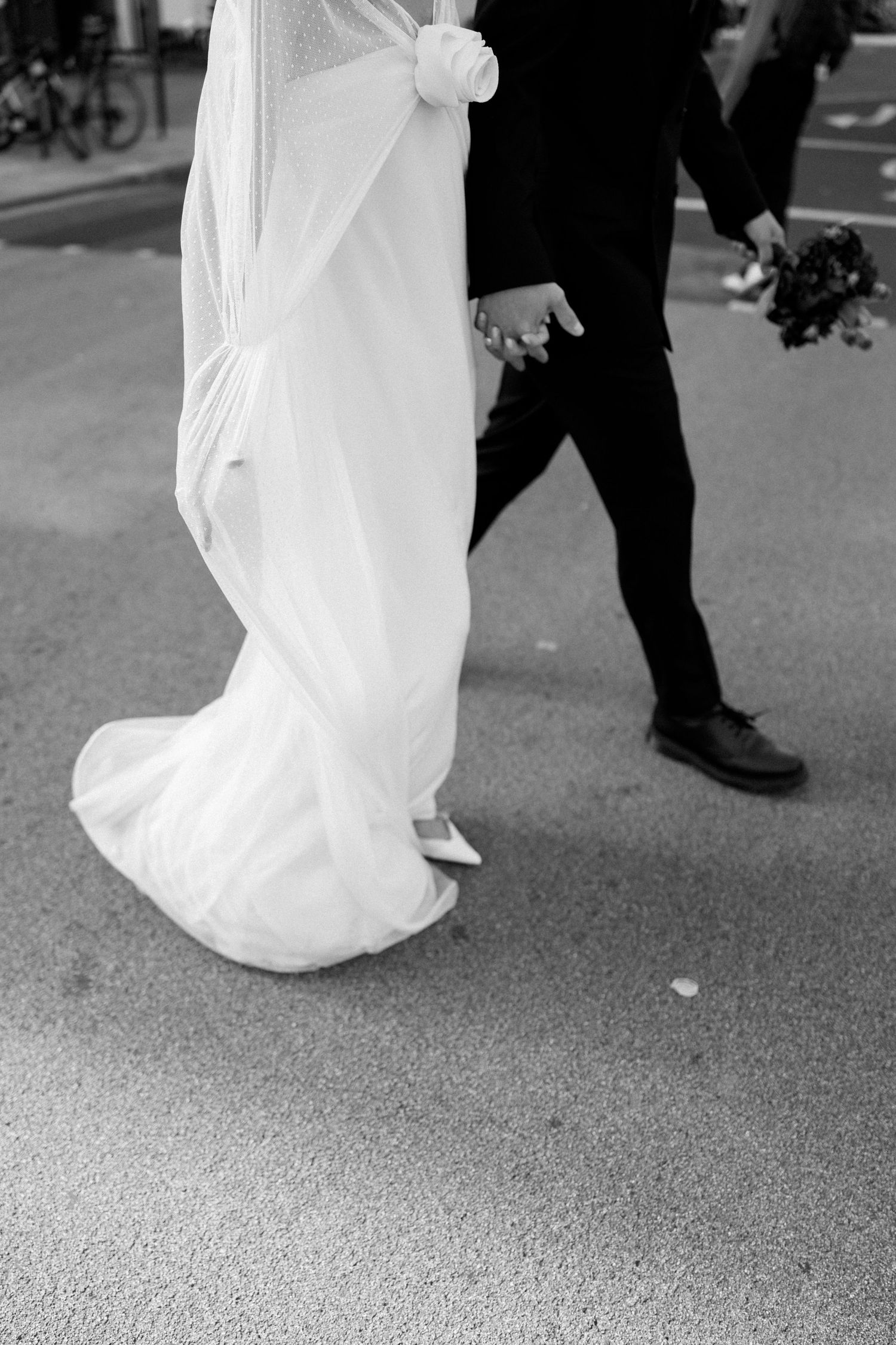 Close-up of flowing white wedding dress and black suit shoes walking on pavement.