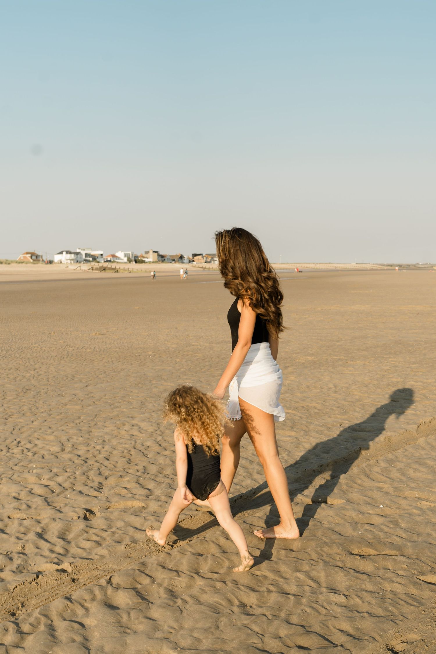 Two people walk together on an empty beach with shadows cast on the sand.