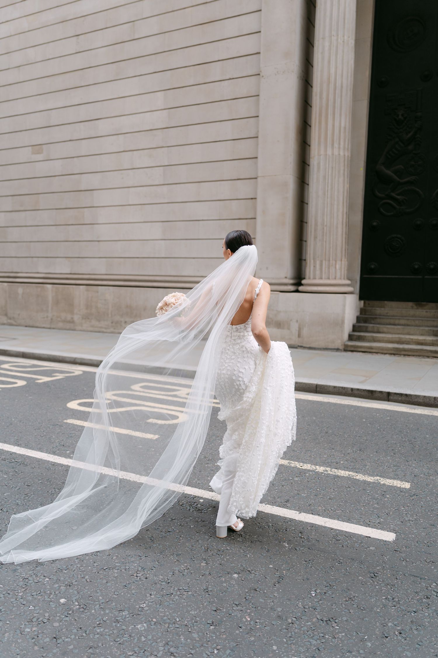 Bride's flowing veil catches wind on city street near classical building columns.