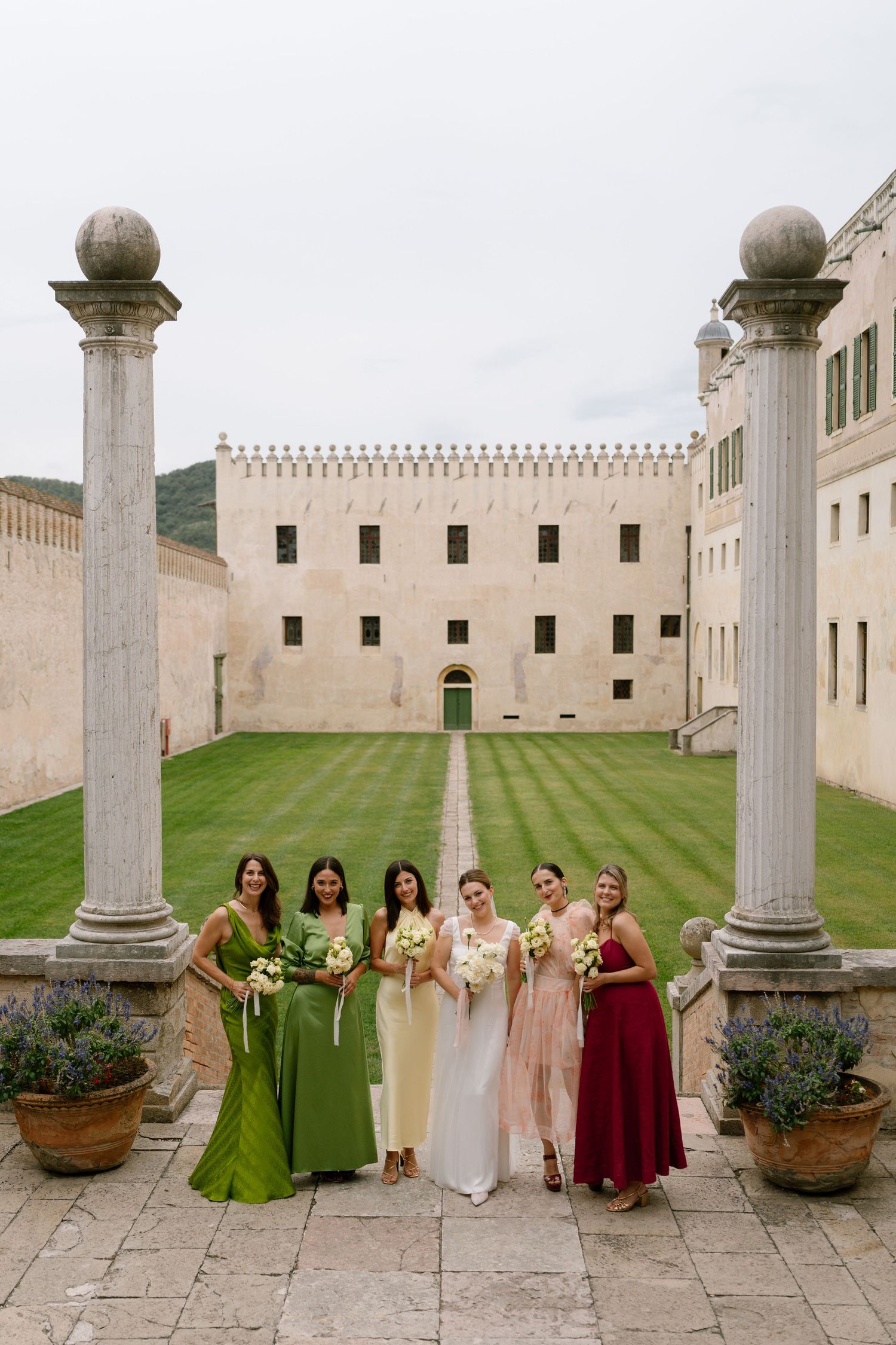Bridal party wearing jewel-toned dresses stands between stone pillars at elegant castle venue.