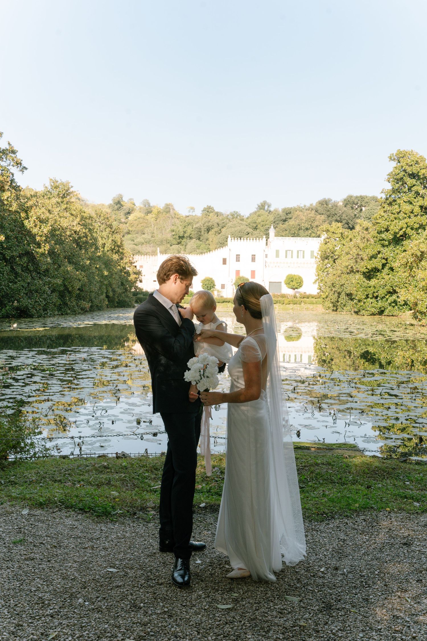 A wedding couple stands by a scenic lake with a white building in the background.