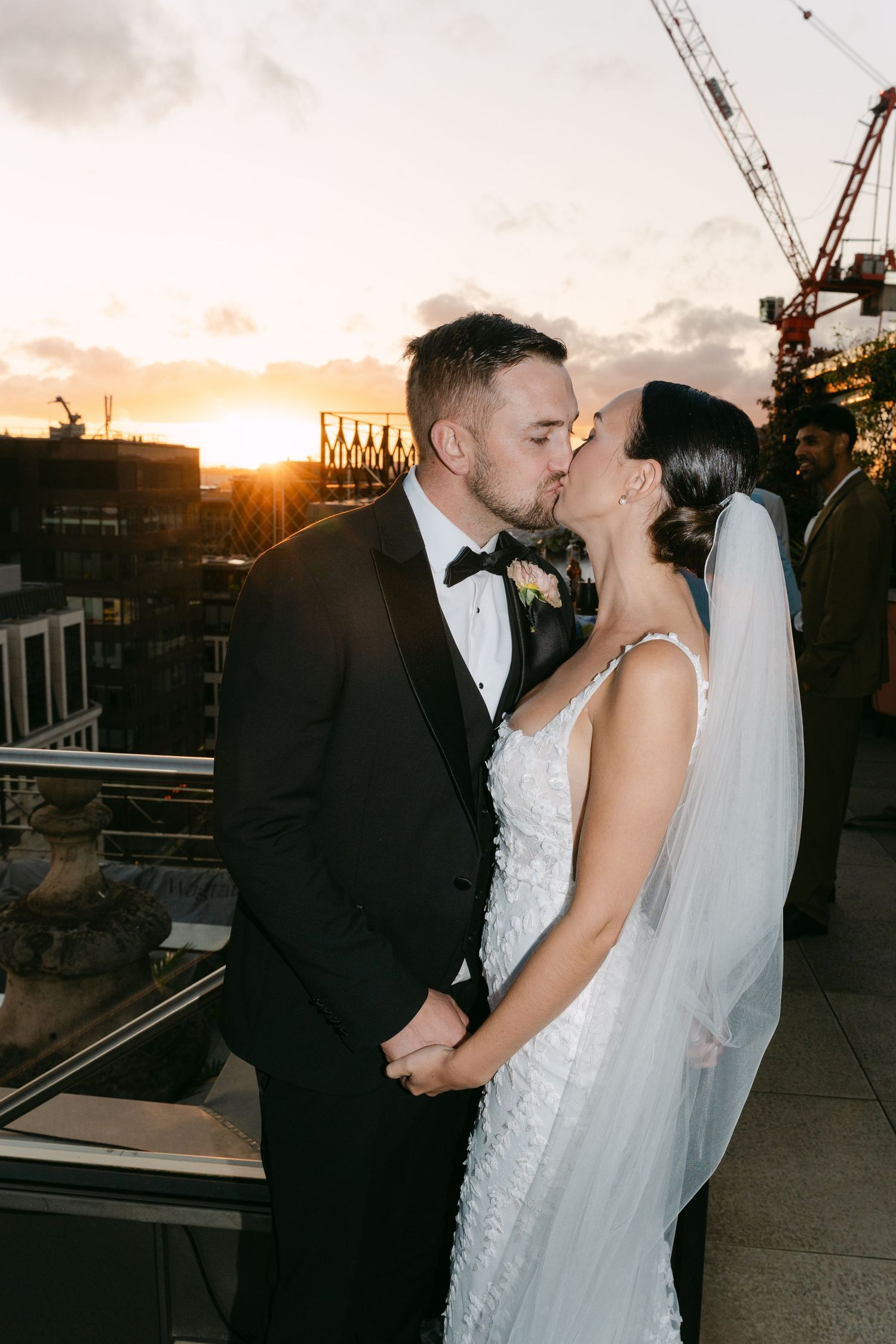 Wedding couple embracing against urban backdrop with construction cranes at sunset.