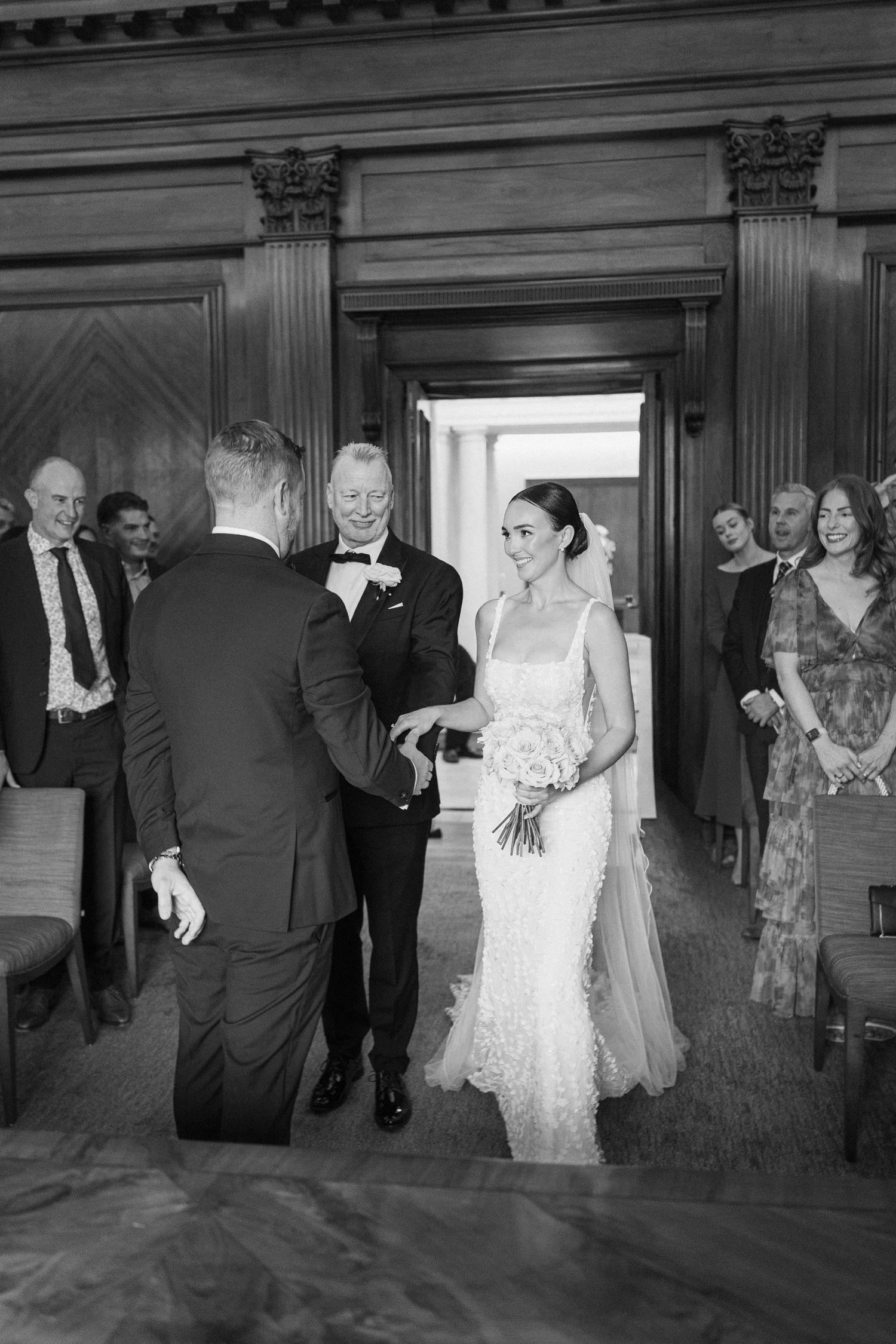 Wedding celebration in a grand wood-paneled hall with guests and wedding party members.