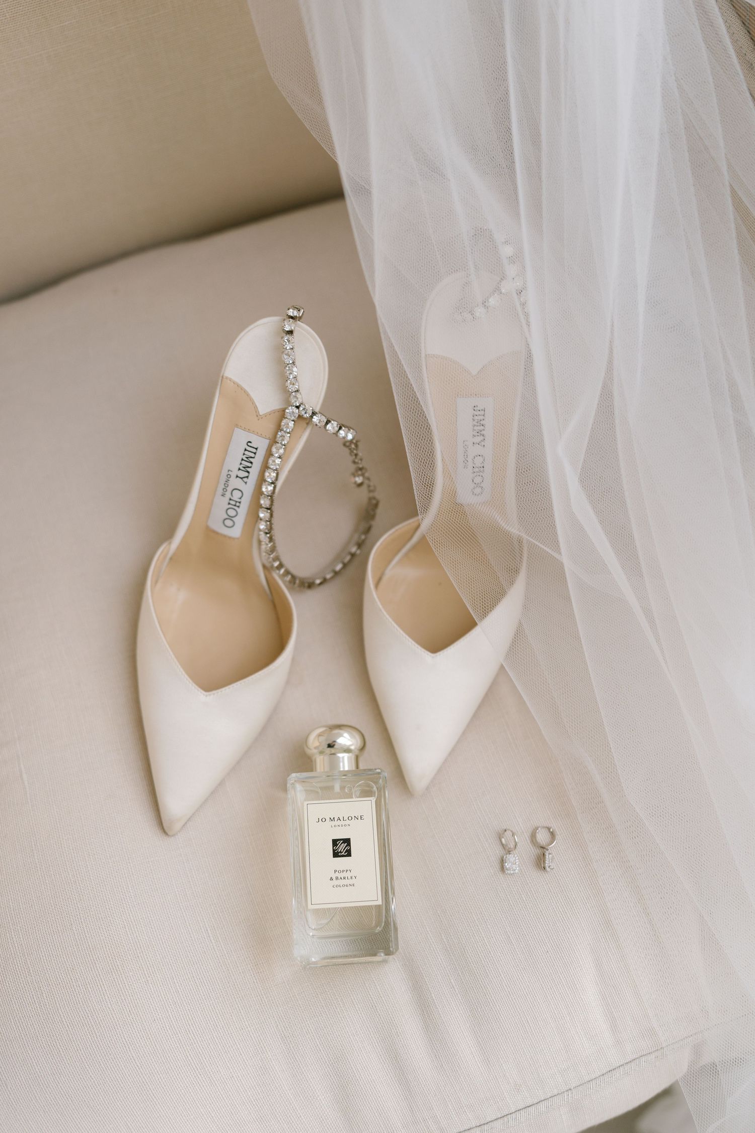Wedding accessories including white shoes, perfume bottle and veil arranged on neutral background.