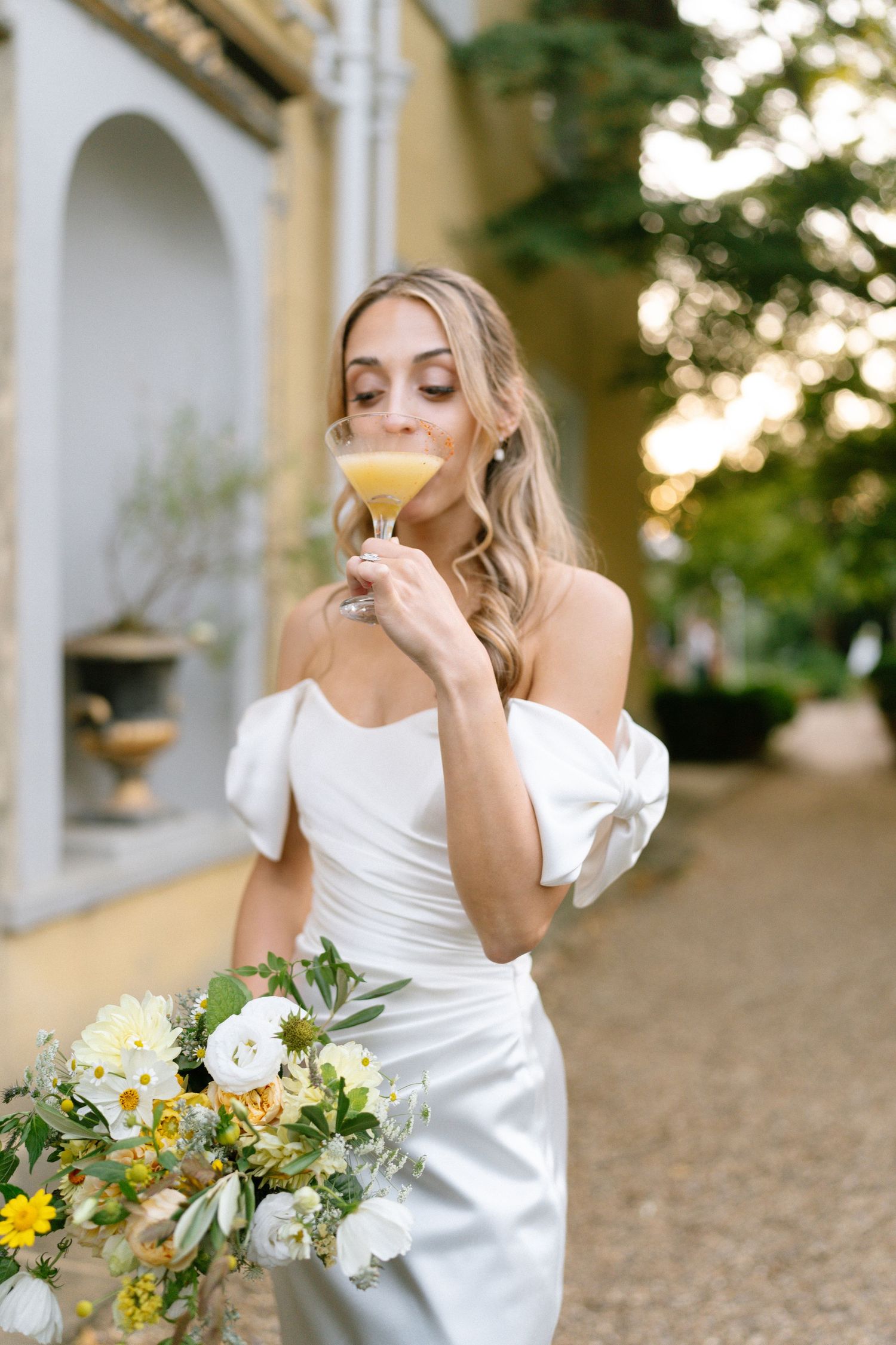 Women in white dresses laugh and hold bouquets against a sunset-lit building exterior.