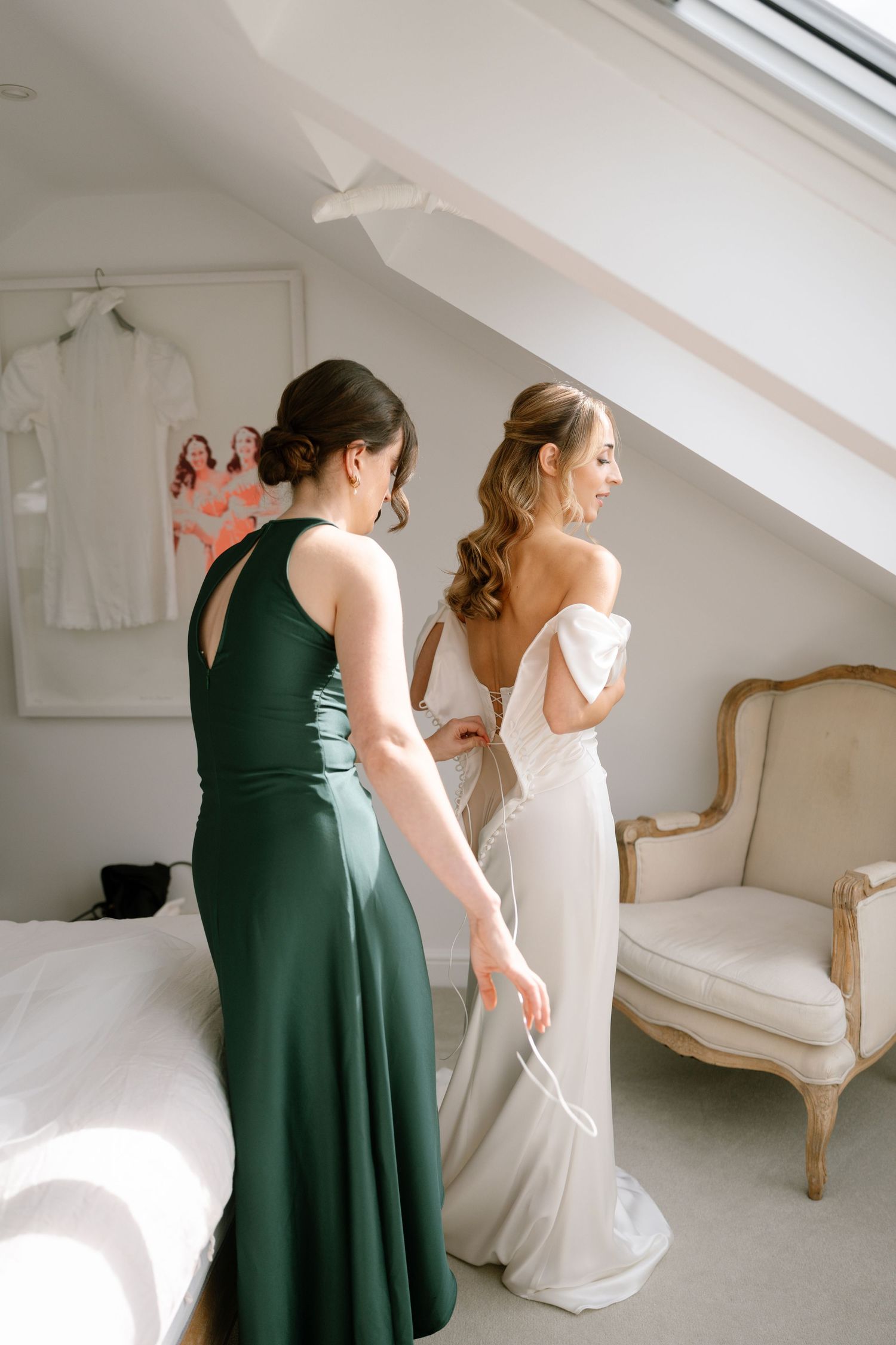 Two women in white and green dresses getting ready in a sunlit room with skylights.