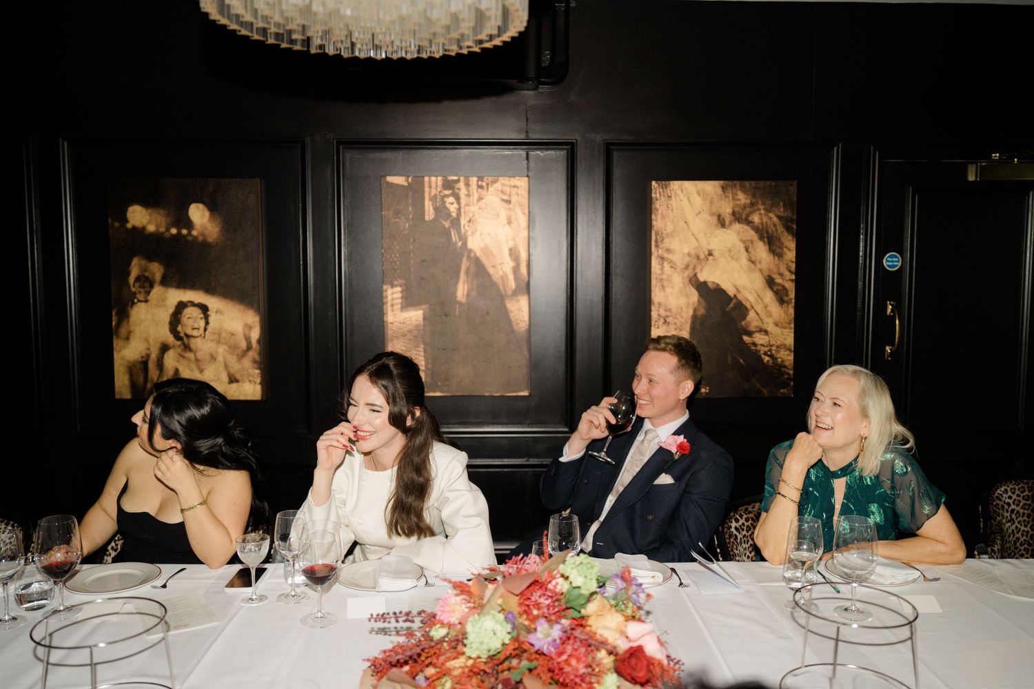 Wedding party guests laugh and celebrate together at a reception dinner table in a dark elegant venue.
