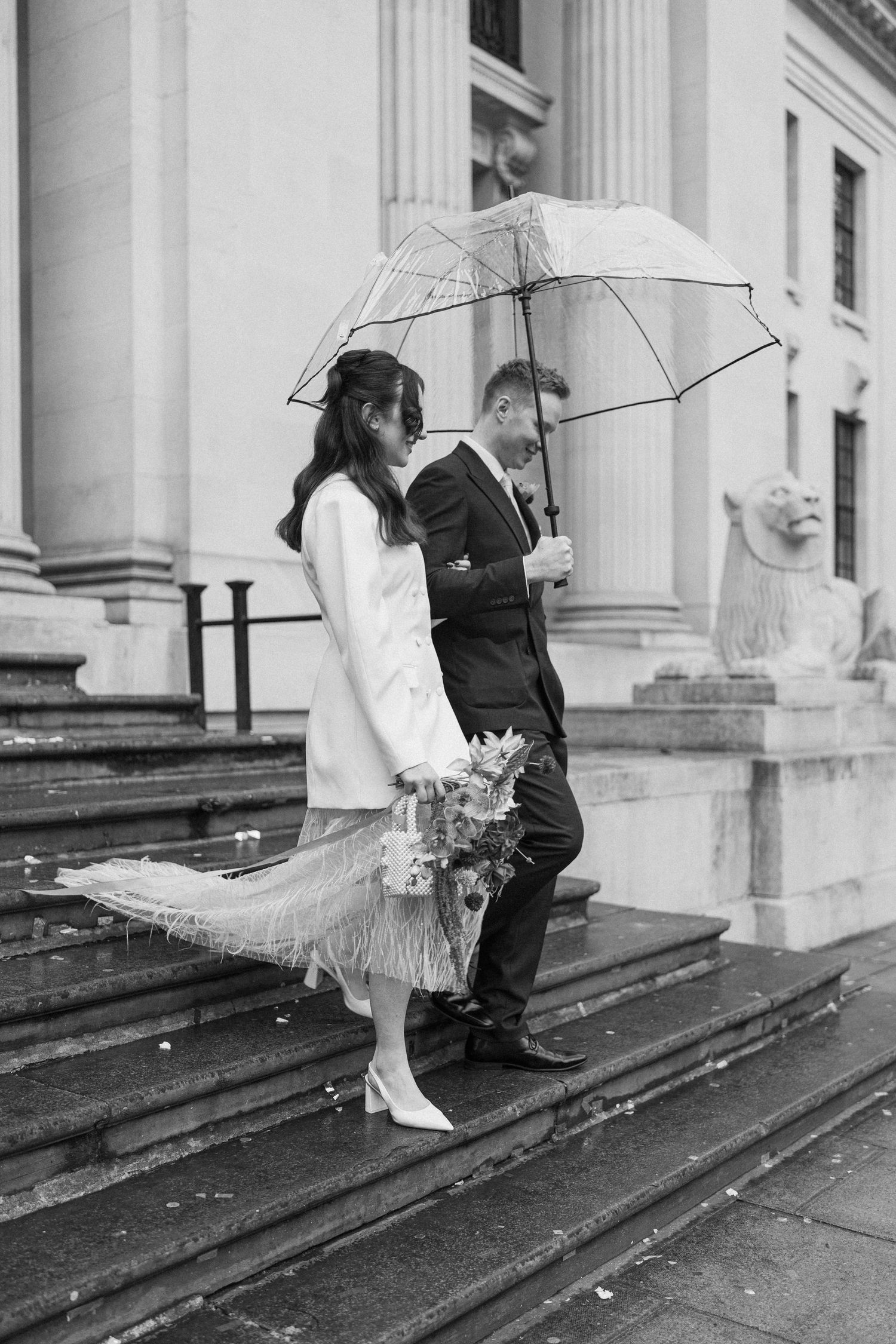 Bride and groom walk down rainy steps sharing a clear umbrella, bride wearing white coat and feather skirt.